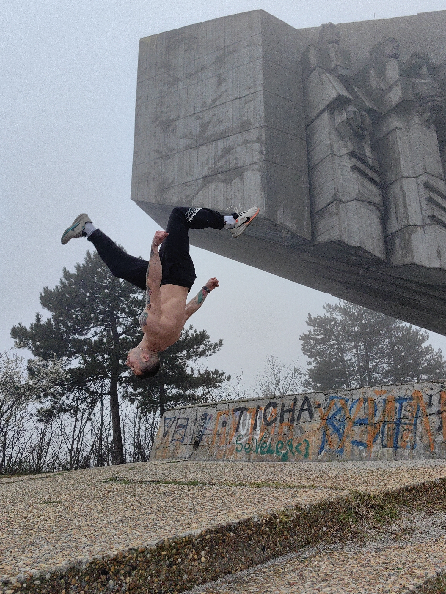 A man is performing a backflip in front of a concrete monument in the city of Varna, Bulgaria. The man has a light complexion, short dark hair, and is shirtless, wearing black pants and athletic shoes. Tattoos are visible on his arms and torso. He is positioned upside down in mid-air, with his legs extended upwards towards the monument. The monument is a large, gray, concrete structure with cubist-style figures carved into its surface. The background is a hazy gray due to fog or overcast weather. Below the monument is a concrete wall with graffiti. Trees are visible behind the wall. The ground is covered with small gravel. The scene appears to be outdoors, possibly in a park or public space.