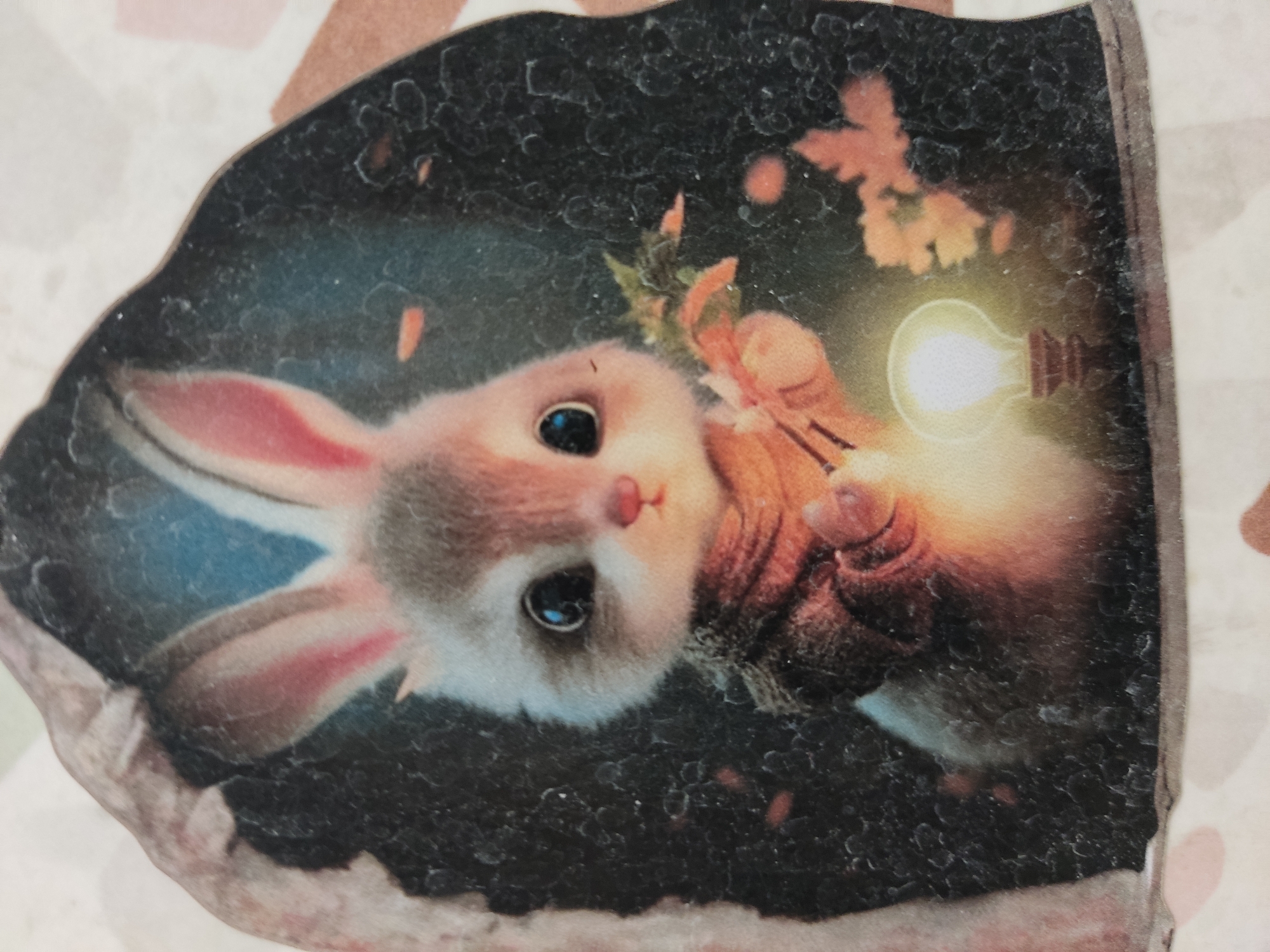 The image is a close-up of a sticker featuring a digitally illustrated rabbit. The rabbit is depicted in a forest setting, holding a glowing lightbulb in its paws. The rabbit has large, dark eyes, pink-lined ears, and is wearing a brown knitted sweater. The background is dark and textured, with hints of leaves and branches. The sticker has a rough, uneven edge, suggesting it might be a handmade or artisanal item. The scene is whimsical and evokes a sense of warmth and curiosity. There is no indication of the city of Noginsk, Russia in this image.