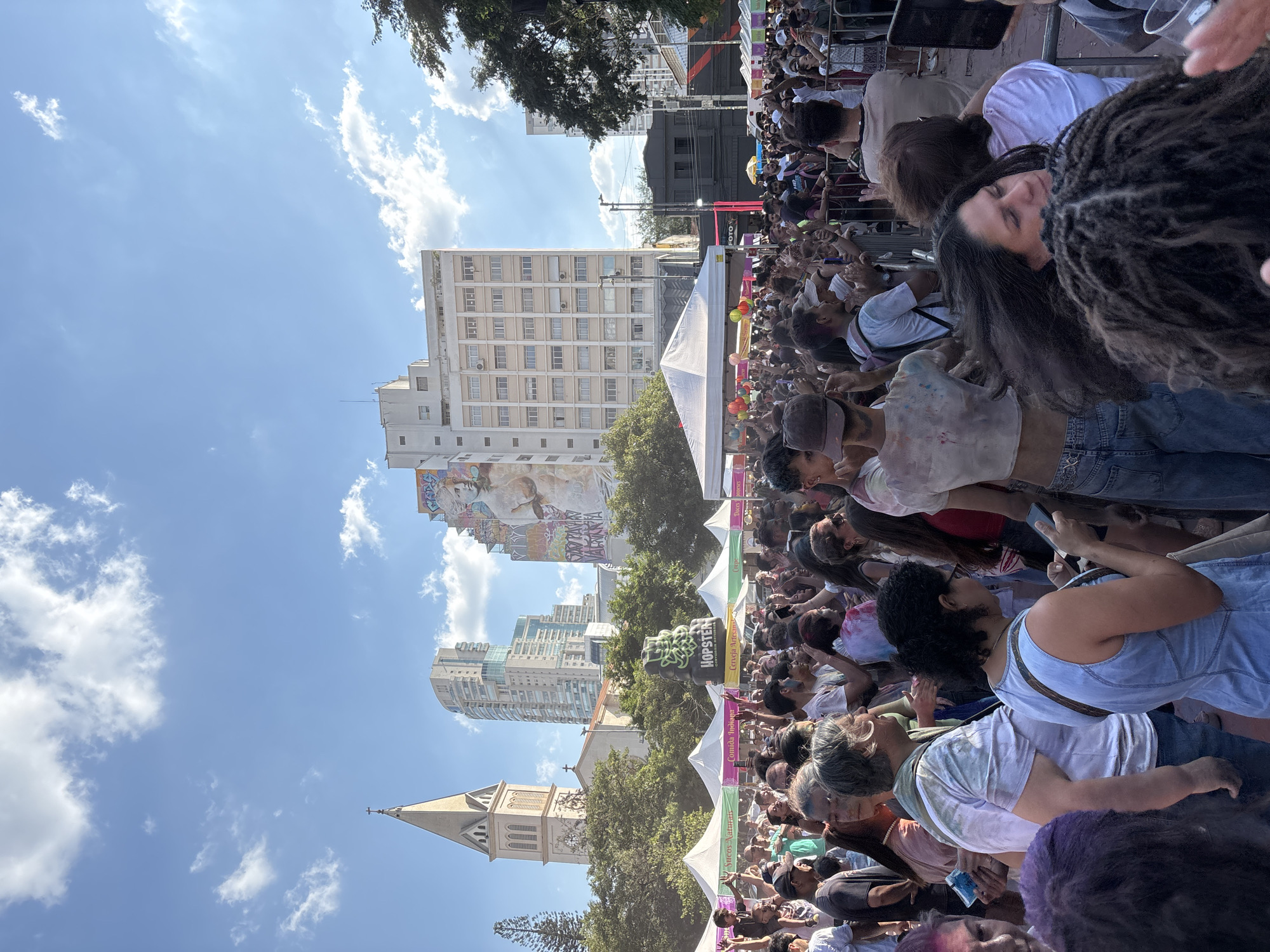 This is a photograph taken outdoors, showing a large crowd of people gathered in what appears to be a public space. The crowd is densely packed, with individuals of various ages and appearances visible.
In the background, there are several tall buildings, indicating an urban environment. One prominent structure is a church steeple with a clock. Banners and signs, including a "HOPSTER" sign, can be seen interspersed among the crowd and the buildings.
The presence of banners and a large gathering of people suggests a public event or celebration. Given the location in São Paulo, Brazil, it is possible that this is a public festival or gathering. The weather appears to be sunny, as indicated by the clear blue sky with scattered clouds.