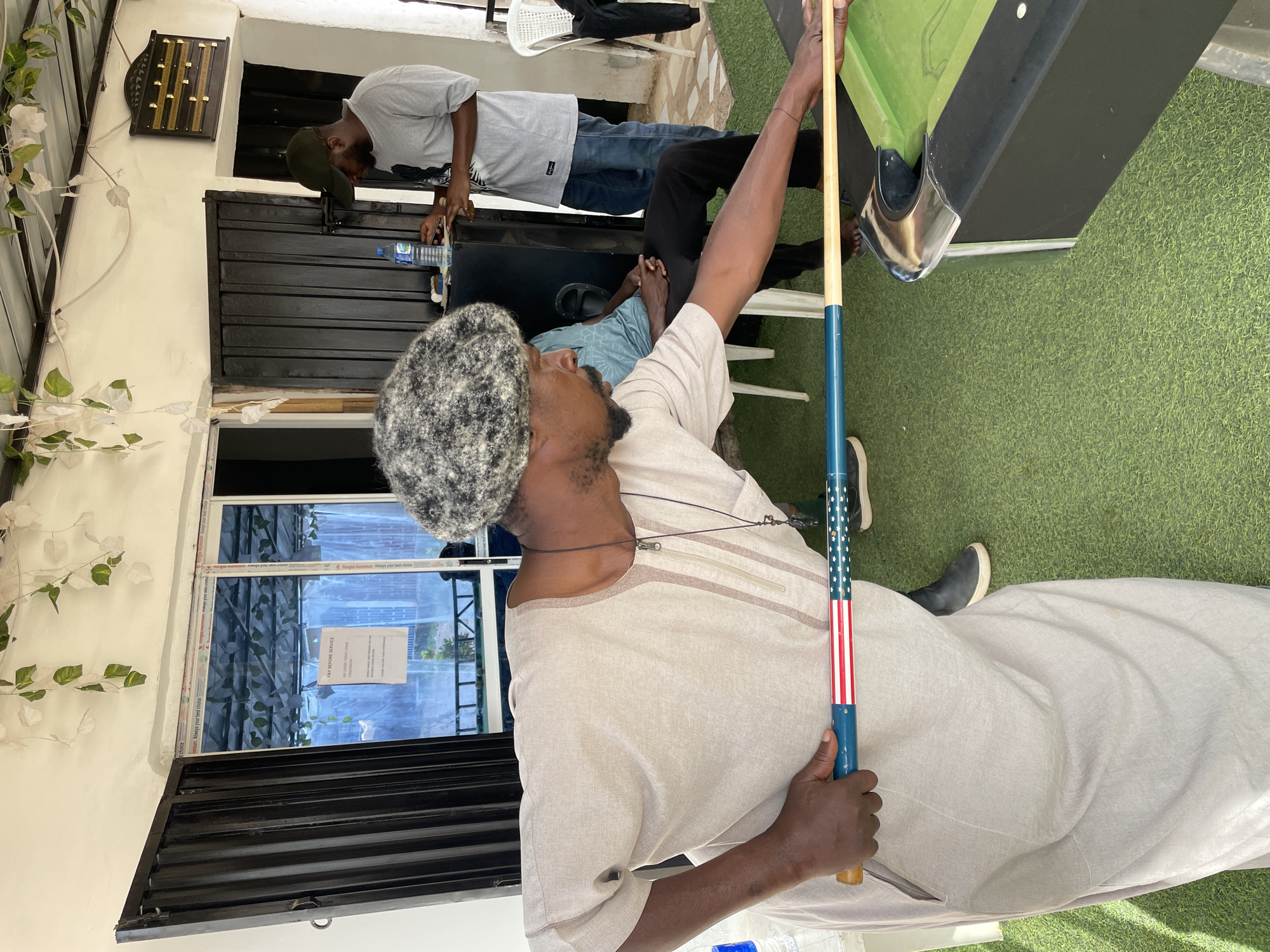 The image depicts a recreational scene in Barakin Akawo, Nigeria, likely an indoor or semi-outdoor game area. In the foreground, a man is seen from the side, wearing a light-colored, long-sleeved traditional garment with a zipped neckline detail and a textured grey and white patterned cap. He holds a blue pool cue adorned with an American flag design (stars and stripes) as he prepares to play pool or is observing the game.
Behind him, another man stands near a black pool table with a green felt surface. This man is dressed in a grey t-shirt and jeans, holding a plastic water bottle labeled "AQUA SAFI" and possibly "NATURAL SPRING WATER". A third person is partially visible, seated in the background. The floor is covered with artificial green turf.
The setting includes white walls, adorned with white climbing plants on the left. On the wall, a wooden scoring board with numbered slots is mounted. Below it, dark, louvered structures are visible. A large window in the background features visible text on a sign that reads "PAY BEFORE SERVICE", "NO CREDIT SALES", "DO NOT DEPOSIT YOUR TRASH HERE", and "THANK YOU FOR YOUR PATRONAGE". The window frame itself shows the text "Triangle Aluminium Always your best choice".
The activity centers around a game of pool, with participants focused on the table. The lighting suggests it is daytime. The overall atmosphere appears casual and relaxed.