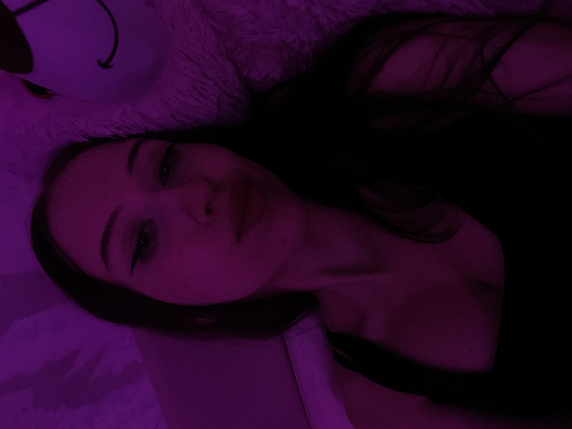 The image is a close-up, low-light selfie taken indoors. A young woman with long, dark hair is the primary subject. She is wearing a black top and has winged eyeliner. The lighting in the scene is predominantly purple, casting a strong hue over everything. To the right of the woman, a portion of a large, fluffy, white object with a cartoonish face is visible. The background appears to be a textured surface, possibly a wall or bedding, also illuminated by the purple light. There is no visible indication of the location being in Votkinsk, Russia.