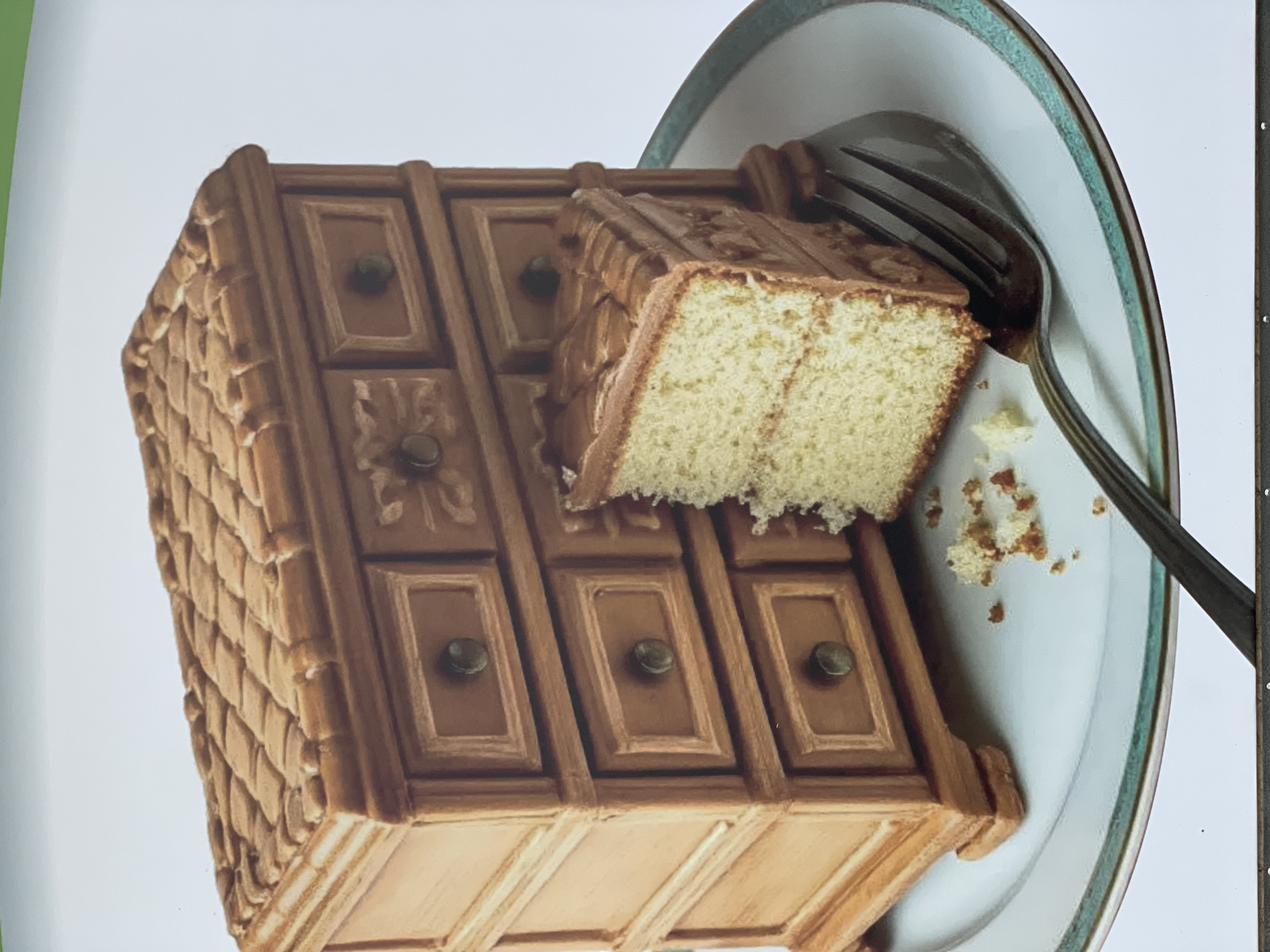 A three-dimensional cake resembling a wooden dresser is positioned on a white plate with a teal rim. A piece of the cake has been cut and sits atop the "dresser," partially removed. A fork rests on the plate next to the cake, suggesting it has been used to cut and possibly eat the dessert. Crumbs are scattered around the cut piece of cake on the plate.
There is no information to indicate that this image has any connection to the city of Zheleznogorsk, Russia.