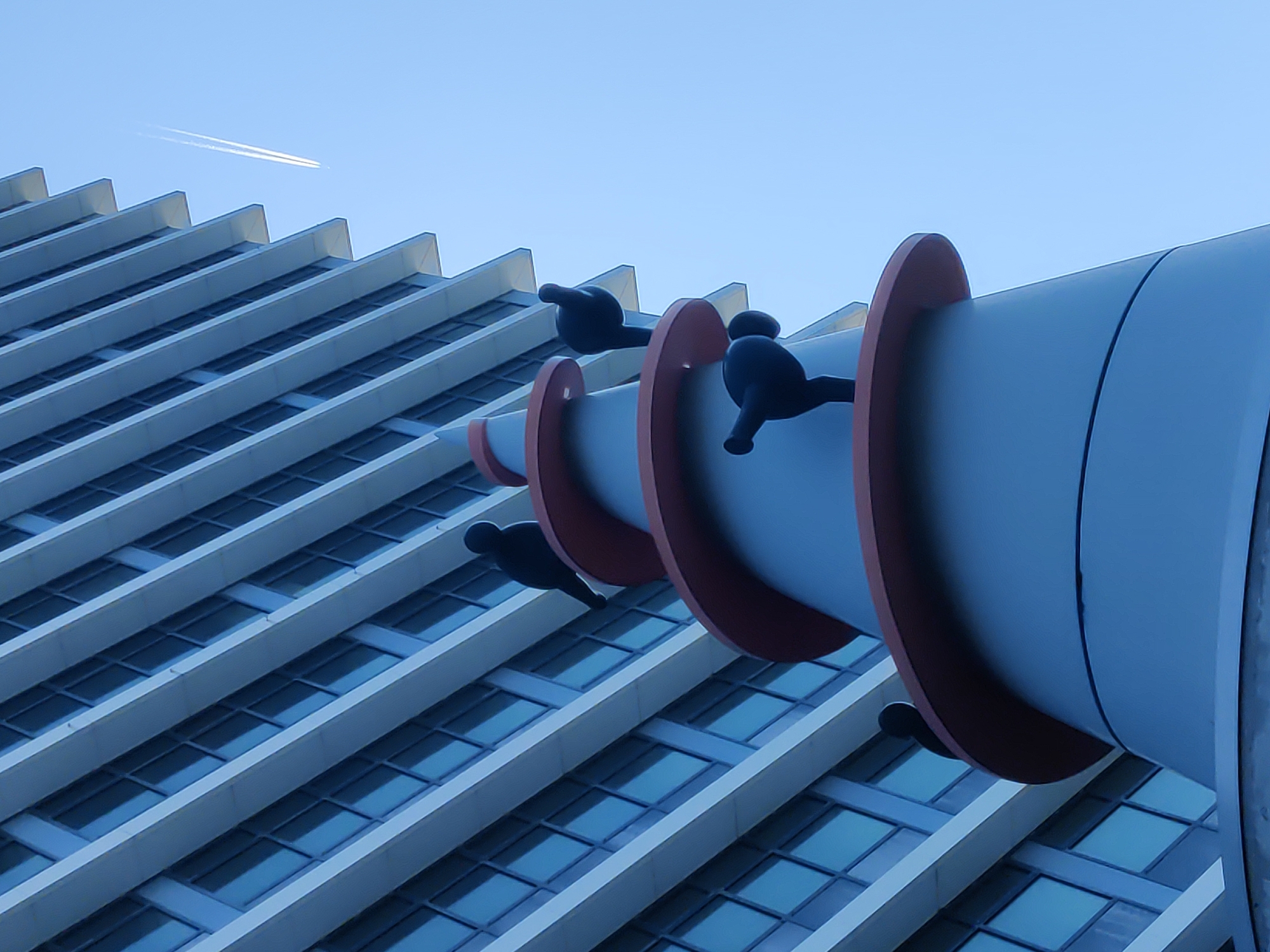 The image captures a low-angle, wide shot of a modern building with a light blue facade and horizontal white accents, possibly a residential or office complex, set against a clear, pale blue sky. The building's structure appears to be a series of stepped levels, creating a sense of depth and architectural interest. In the foreground, a whimsical, sculptural element is visible. It is a tall, conical structure in muted grey and red, adorned with several stylized, black, bird-like figures. These figures are in various poses, some appearing to perch or stand on the red bands that encircle the conical structure. The sculptures have a minimalist, cartoonish aesthetic. High in the sky, a contrail from an airplane streaks across the scene, adding a dynamic element to the otherwise still composition. The lighting suggests it is daytime, with the sun casting subtle shadows that highlight the architectural features. The overall impression is one of a blend of modern urban architecture and playful public art. This image is located in the city of Rijswijk, Netherlands, specifically at the former headquarters of the Dutch Ministry of Social Affairs and Employment, now known as the Ministry of Social Affairs and Employment and the Ministry of Health, Welfare and Sport.