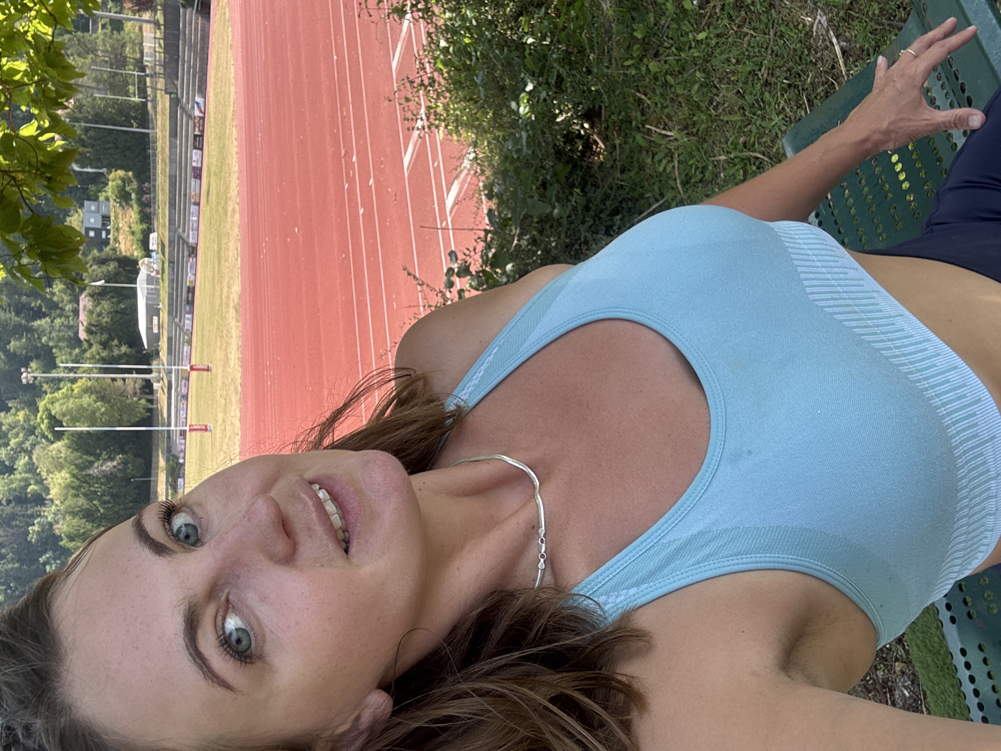 A woman is taking a selfie outdoors. She is wearing a light blue sports bra and dark blue leggings. She has light brown hair and blue eyes. She is smiling and looking at the camera. The background is a red running track with green grass and trees. There are stadium bleachers in the distance. The scene appears to be a sports field in the city of Saint-Claude, France.