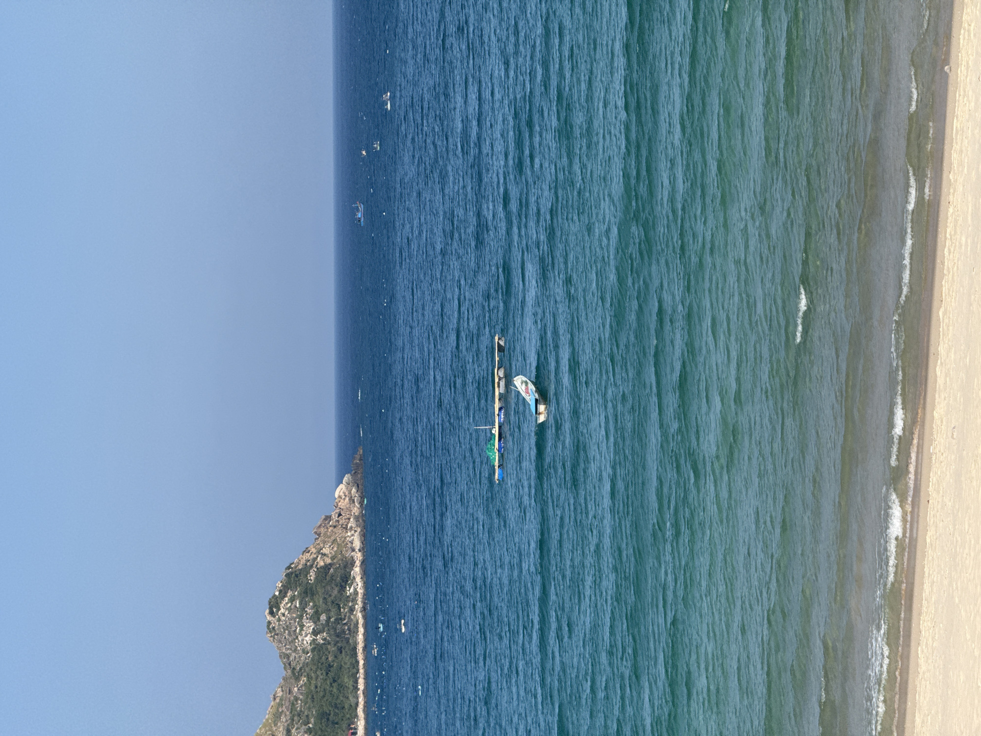 The image depicts a serene coastal scene under a clear blue sky. In the foreground, a sandy beach meets the gentle waves of the ocean, with the water transitioning from a lighter turquoise near the shore to a deep blue further out. Several boats are scattered across the water. Two small white boats are visible, one closer to the foreground and another further away. A larger, flat raft-like structure with a net and some blue containers is also present. In the mid-ground, a rocky, green-covered island juts out of the sea, with a few scattered buildings visible on its slope. The horizon shows a few more distant boats, suggesting ongoing maritime activity. The overall impression is one of tranquility and a typical day at a seaside location.