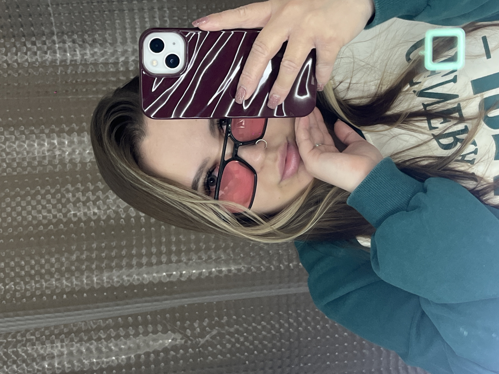 An indoor mirror selfie features a young person, likely female, in Krasnodar, Russia, holding a smartphone to capture their reflection. The person has long, multi-toned hair, primarily brown with blonde highlights framing the face. They are wearing black-framed glasses with distinct pink-tinted lenses, a small ring on their right hand, and a nose ring. Their fingernails are manicured with a light pink, possibly glittery, polish.
The individual is dressed in a dark teal or dark green long-sleeved top or hoodie, worn over a lighter-colored t-shirt. Part of a dark, blocky text is visible on the t-shirt, reading "A M E" (possibly part of "GAMER" or "AMERICAN"). A small, light green, square-shaped accessory or clip is present on the t-shirt or in the hair near the upper right side of the head.
The smartphone, with visible dual camera lenses, is encased in a maroon or dark red protective cover featuring white, wavy, diagonal stripes. The person holds the phone up, positioning it to take the photo. The background is a textured, light-colored surface, appearing to be a reflection of something like bubble wrap or a patterned wall, indicating an indoor setting. The person's expression is neutral, and they are looking directly into the camera's reflection.