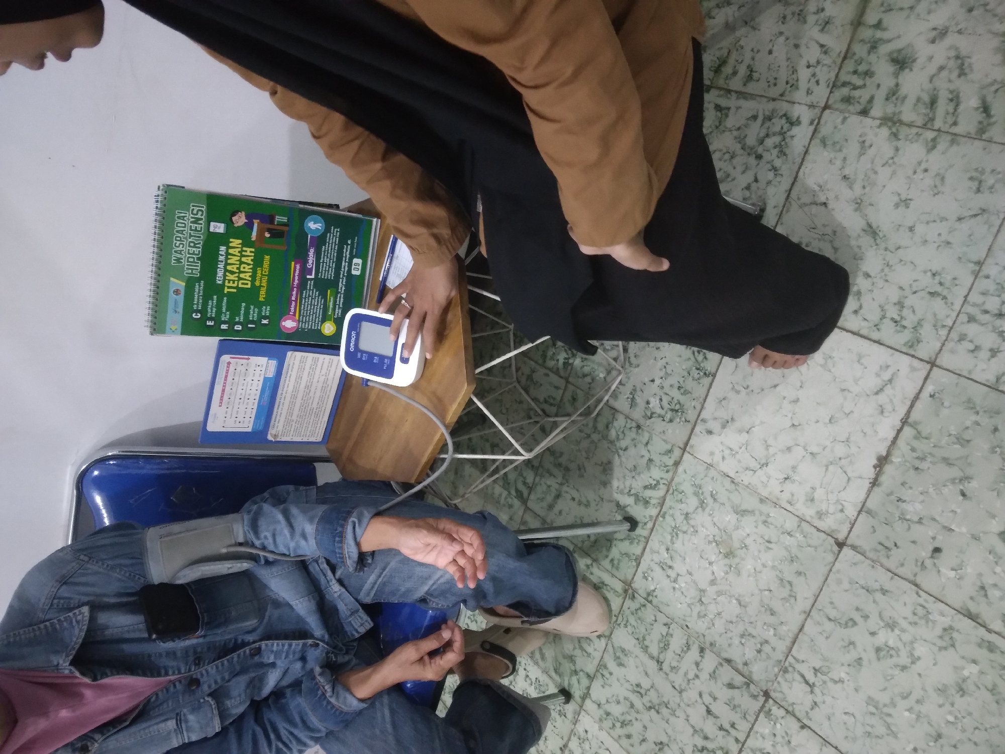 The image depicts an indoor scene where a person is having their blood pressure taken. The person being measured is wearing a denim jacket over a pink shirt and blue jeans, with their left arm fitted with a blood pressure cuff. They are seated in a blue chair. To the right, a person wearing a brown top and a black headscarf is partially visible, operating a white Omron blood pressure monitor. A small wooden table is positioned between them, holding the blood pressure monitor and a spiral-bound booklet. The booklet features text in Indonesian, with the title "Waspadai Hipertensi" (Beware of Hypertension) prominently displayed. The background shows a light-colored wall and a patterned tiled floor. The overall setting appears to be a health clinic or a similar public health facility.