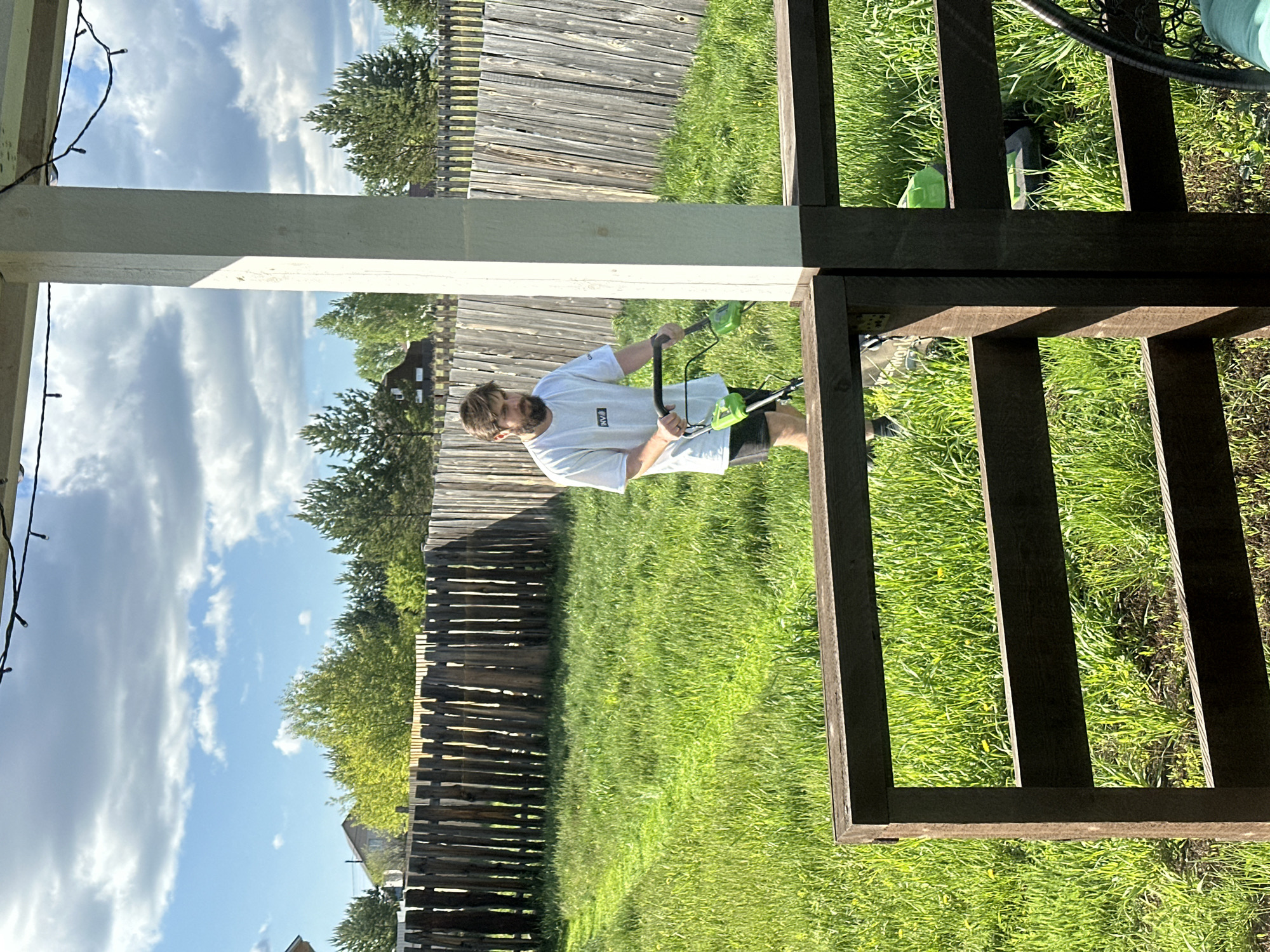 This image depicts an outdoor scene in a backyard, likely in Kuda, Russia, captured during bright daylight under a partly cloudy sky. The main subject is a man with a beard, wearing a white t-shirt with a "P.A.M." logo on the left chest, and dark shorts. He is actively engaged in trimming grass using a bright green and black electric string trimmer.
The man is positioned beneath a wooden structure, possibly a porch or deck, which features prominently in the foreground and frames the scene. A white wooden support post is visible on the left, adorned with string lights. Dark brown wooden railings and framing of the structure are seen on the right and across the bottom of the image. The ground is covered in lush green grass, suggesting a well-maintained or recently overgrown lawn. In the background, a rustic wooden fence stretches across the width of the yard, with various trees and a glimpse of a building visible beyond it. The clear shadows indicate a sunny day, providing strong contrast to the bright green grass and blue sky. The man appears focused on his task.