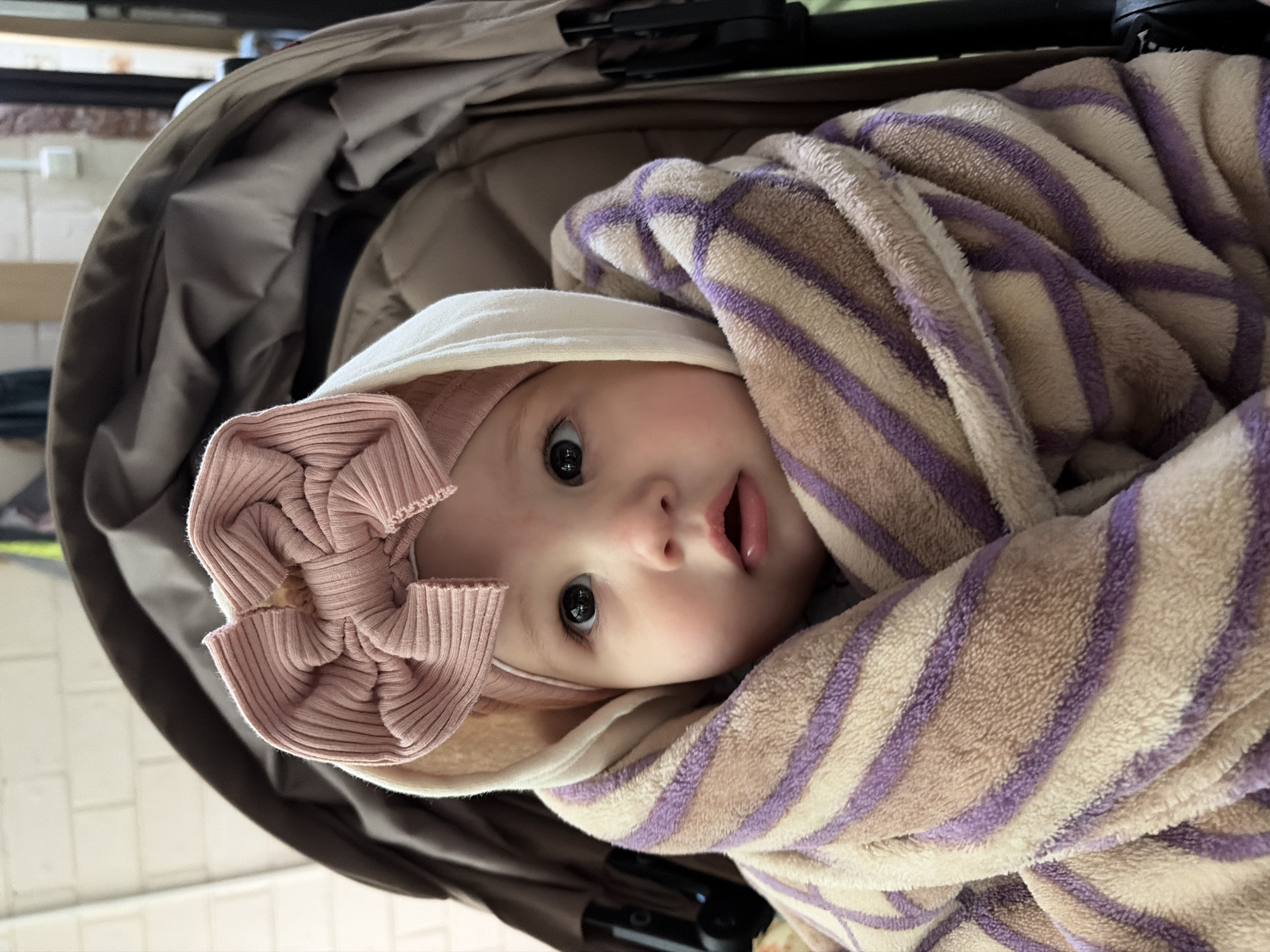 A baby with fair skin and dark eyes, wearing a pink ribbed headband with a bow and a white hooded undershirt, is wrapped in a striped beige and purple blanket. The baby is lying in a tan-colored stroller. The location of Novopokrovskaya, Russia, cannot be determined from this image.