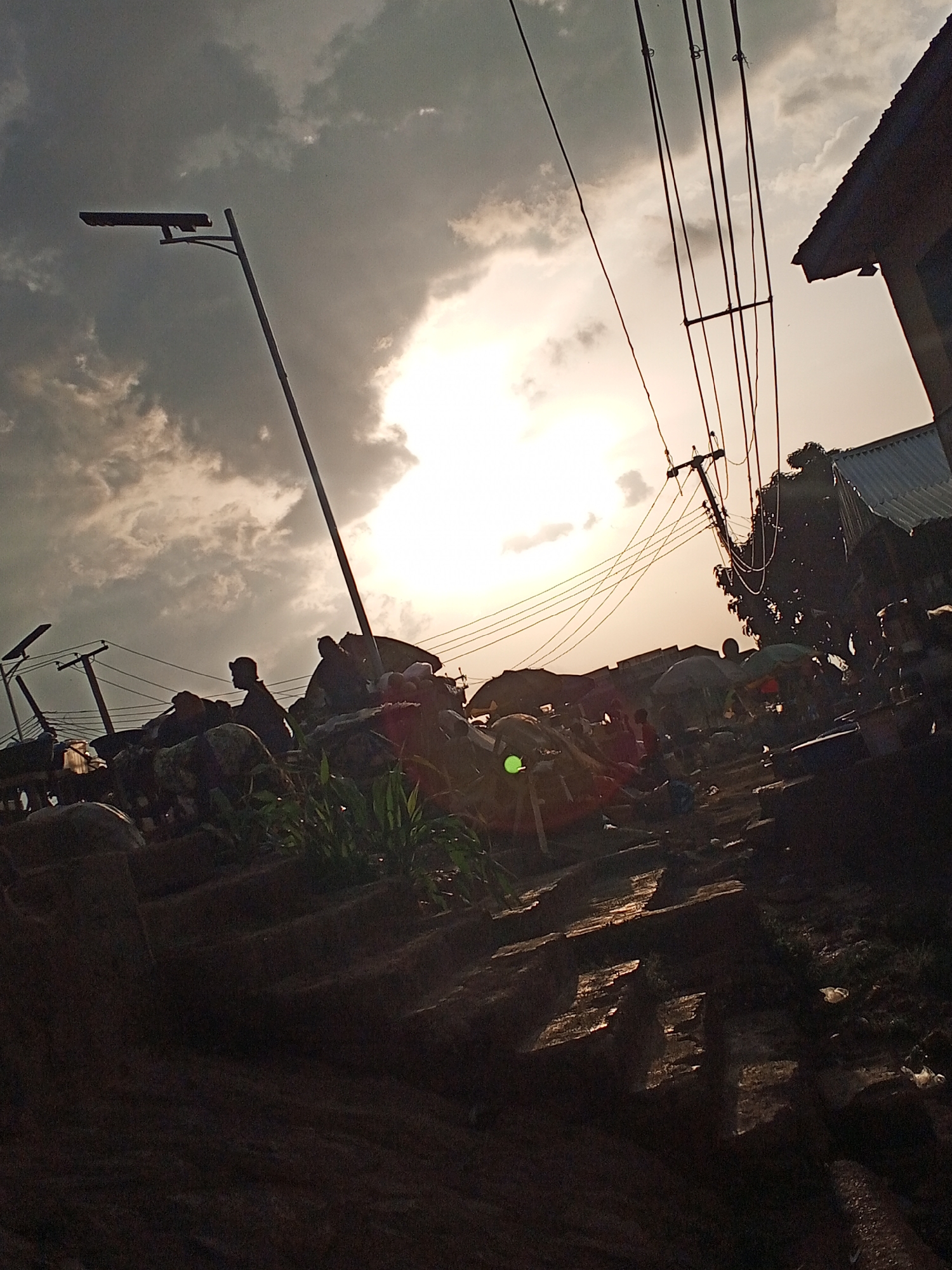 The image depicts an outdoor scene in Gwong, Nigeria, captured at dusk or dawn. The bright sun shines through heavy clouds, casting long shadows and creating high contrast. In the foreground, there are steps or a rugged terrain, possibly made of stone or earth.
A street market scene is visible with people and goods under various umbrellas and makeshift canopies. Silhouetted figures of people are visible throughout the scene. Utility poles and power lines crisscross the sky. A modern street lamp is also visible. The scene conveys the bustling activity of a marketplace at a time of day with dramatic lighting conditions.