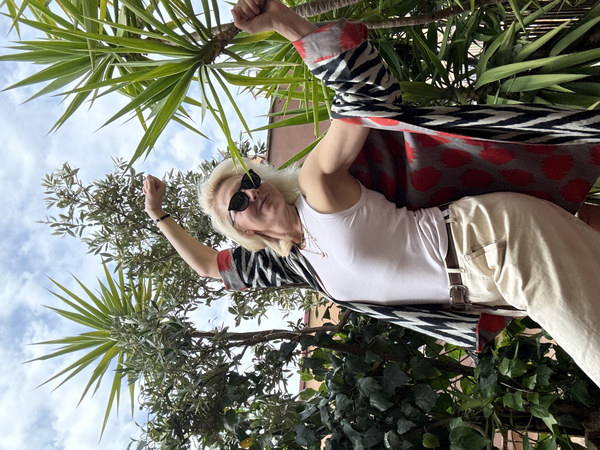 A person stands outdoors, posing with arms raised, amidst foliage and under a partially cloudy sky. The individual wears sunglasses, a white tank top, light-colored pants with a brown belt, and a patterned jacket with red and gray elements. The surroundings include trees with spiky leaves, leafy green plants, and a glimpse of a building behind the vegetation. The location is suggested to be in Rome, Italy.
