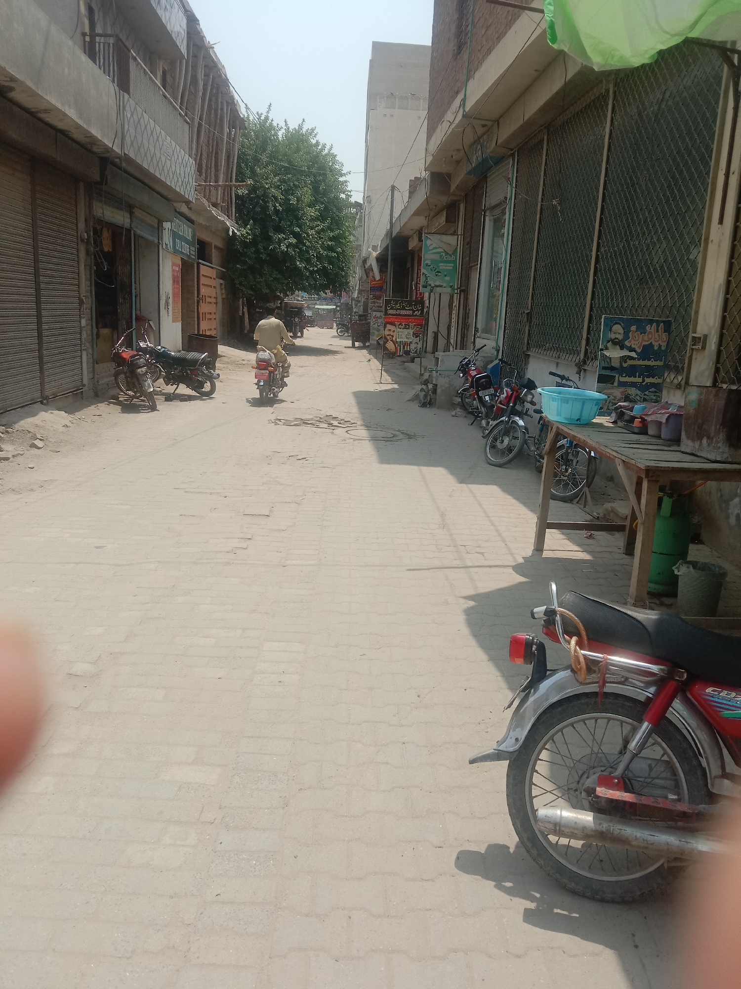 The image depicts a street scene in Depalpur, Pakistan. The street is paved with brick-like stones and appears to be moderately trafficked. On the left side of the street, buildings with shuttered storefronts line the road. One storefront displays an advertisement with text in Urdu script and images of men. Further down the street, a tree provides shade. On the right side of the street, more storefronts are visible, some with metal grates. A wooden table is set up outside one of these establishments, with a blue plastic basin, some containers, and a green gas cylinder beneath it. Several motorcycles are parked along the street. One man on a motorcycle is riding away from the camera, wearing a light-colored shirt and trousers. Another motorcycle is parked in the foreground on the right, showing its rear wheel, fender, and exhaust pipe. The sky is clear and the overall lighting suggests daytime. The environment appears to be a commercial area within a Pakistani city.
