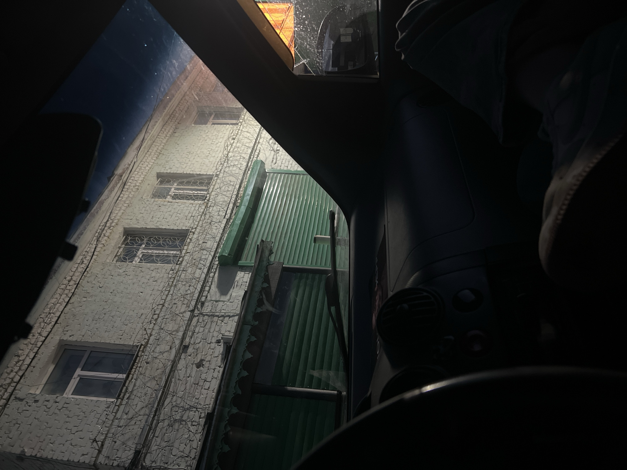 The image is taken from inside a car, looking out at a building at night. The building is made of white brick and has several windows. There is barbed wire along the top of the building and across the windows. Below the windows are green roller shutters. The interior of the car is dark, with only the dashboard and the steering wheel visible. The scene appears to be a street in the city of Prudys, Russia.