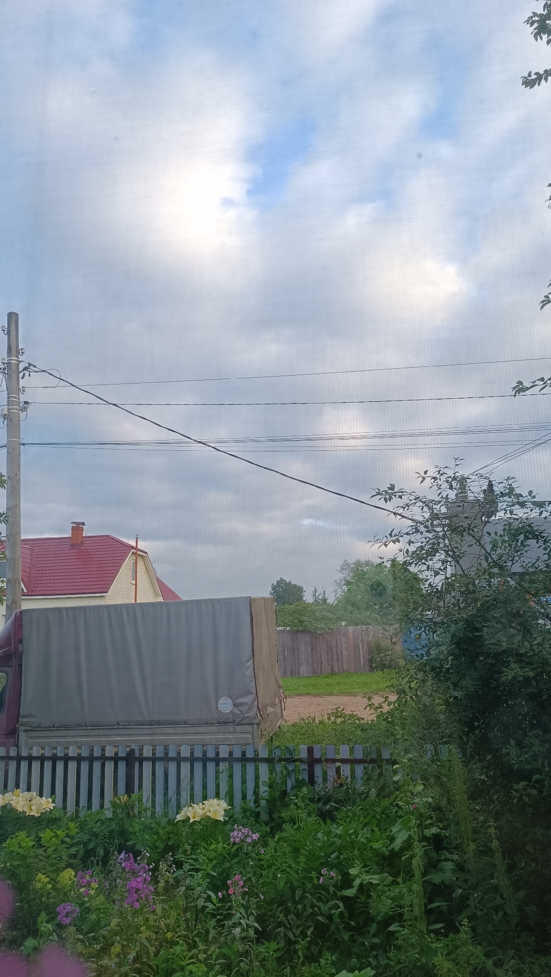 The image depicts a rural scene, possibly in Karsha, Russia, characterized by a cloudy sky and a residential area. A light-colored house with a red roof and a chimney is visible behind a metal fence. A truck with a gray tarp-covered cargo area is parked in front of the fence. Lush green vegetation and flowers are growing in the foreground. Electrical wires are strung across the sky, supported by a wooden pole. In the background, a wooden fence separates the property from a grassy area. Trees and foliage complete the landscape. There are no people or animals present in the image.
