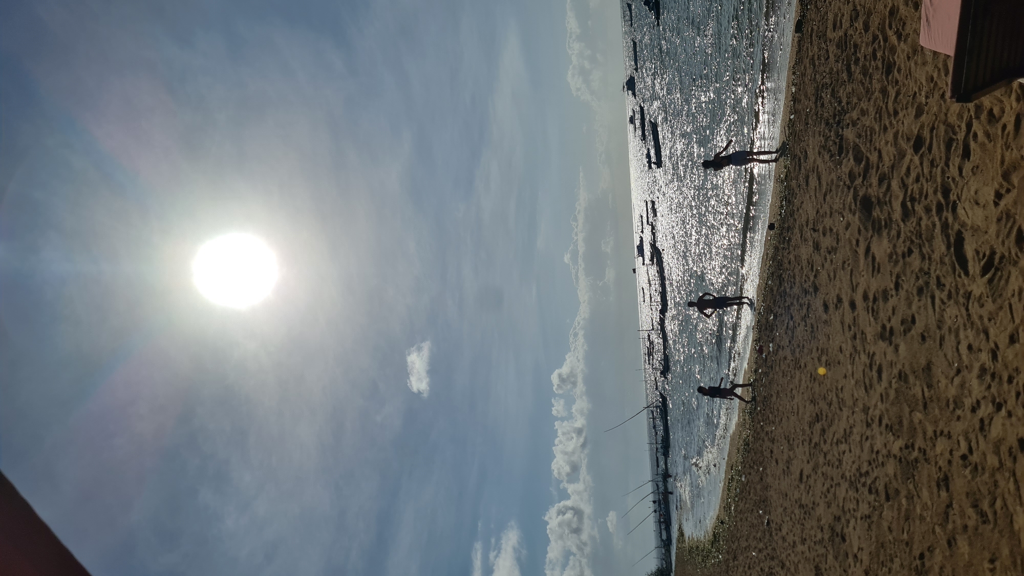 The image depicts a sun-drenched beach scene, likely in Denpasar, Indonesia, characterized by the bright, overexposed sun dominating the upper portion of the frame. Below the sun, a hazy blue sky with scattered clouds transitions to the shimmering, sunlit ocean. Numerous small boats, appearing as silhouettes against the glare, are anchored or drifting on the water, with their tall masts visible. The shoreline is a sandy beach, with footprints and some scattered debris. Three individuals are present on the wet sand near the water's edge. One person, a child, is walking along the shoreline, while two others, likely teenagers or young adults, stand and face the water. The overall atmosphere is one of a bright, possibly hot, day at the coast.