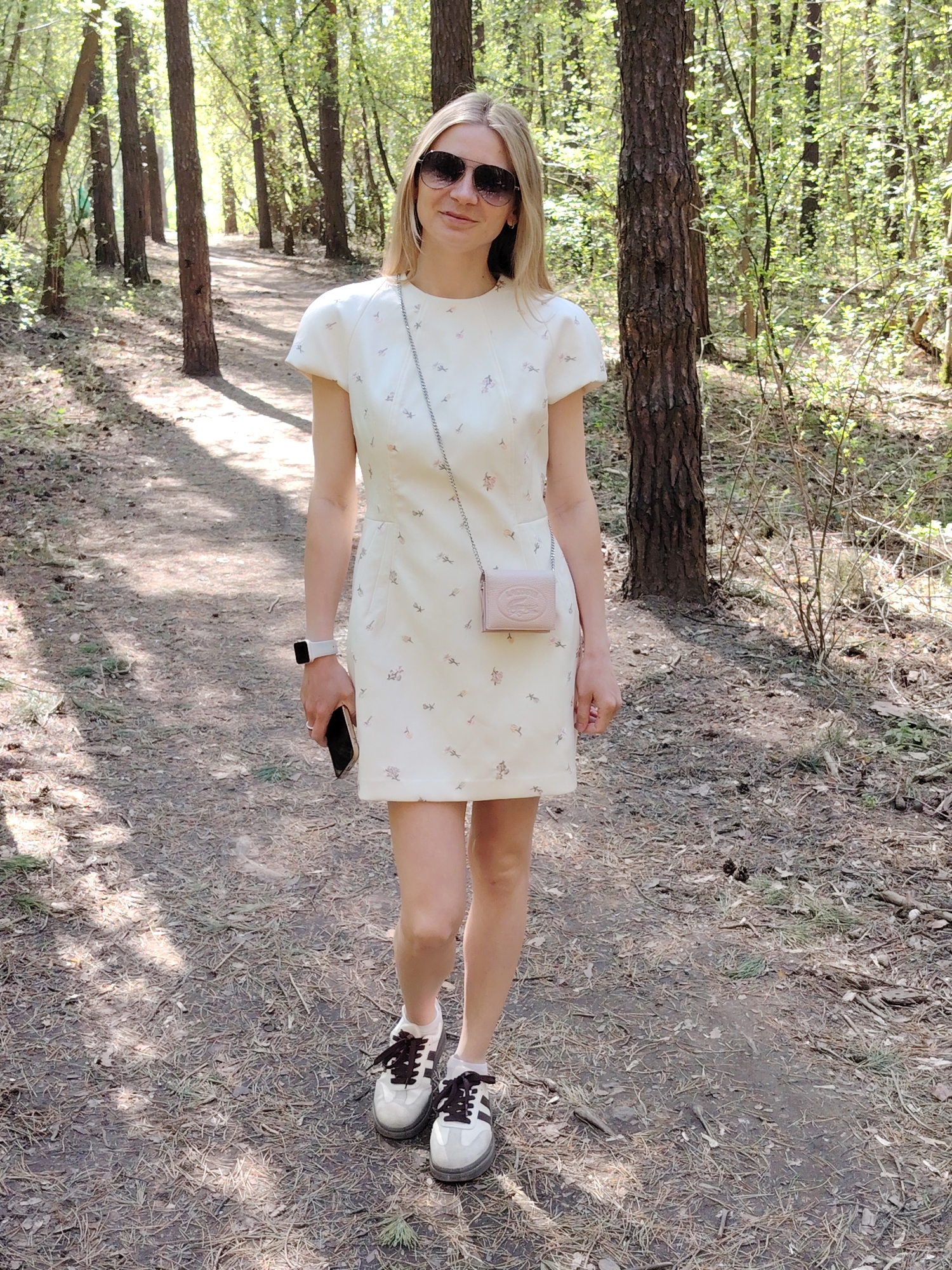 A woman stands on a path in a forest. She is wearing a light-colored dress with a floral pattern, sunglasses, and white sneakers with brown laces. She carries a small, light pink purse with a chain strap and holds a smartphone in her left hand, on which is a smartwatch. The path is covered with fallen leaves and needles, and the forest consists of tall trees with green foliage. The scene is brightly lit, suggesting it is daytime. The location is Chaikovsky, Russia.