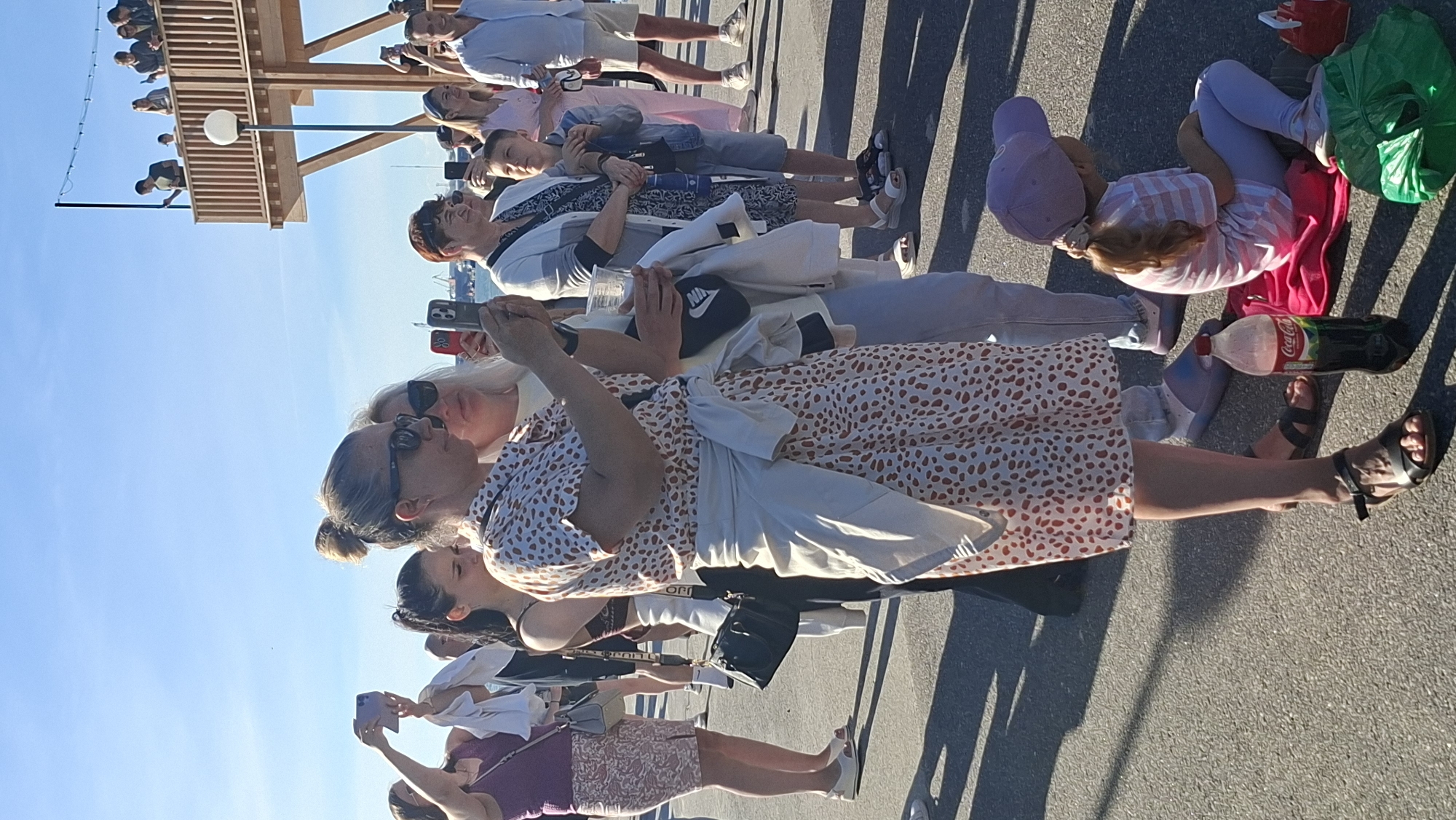 A group of people are gathered outdoors on a sunny day, possibly at a waterfront or promenade, given the presence of a pier-like structure in the background and what appears to be a harbor with ships in the distance. The clear blue sky suggests good weather. The people, of varying ages, are dressed casually. Some are standing, while others are seated or leaning against a railing. Several individuals are holding up their mobile phones, appearing to take pictures or videos of something out of frame, or perhaps of each other.
In the foreground, a woman in a patterned dress is prominently featured, her hands clasped in front of her. To her right, another woman with blonde hair and sunglasses is looking towards the camera. Further down, a child in a pink and white striped shirt and a purple hat is sitting on the ground, with a green bag and a large bottle of Coca-Cola beside her. Another person in a white top and light-colored pants is also seated.
The scene suggests a relaxed, possibly tourist-oriented atmosphere. The bright sunlight and clear sky indicate it is daytime, likely midday or afternoon. The visible text includes "Coca-Cola" on the beverage bottle and a "NIKE" logo on a black baseball cap. The overall impression is one of people enjoying an outdoor excursion.