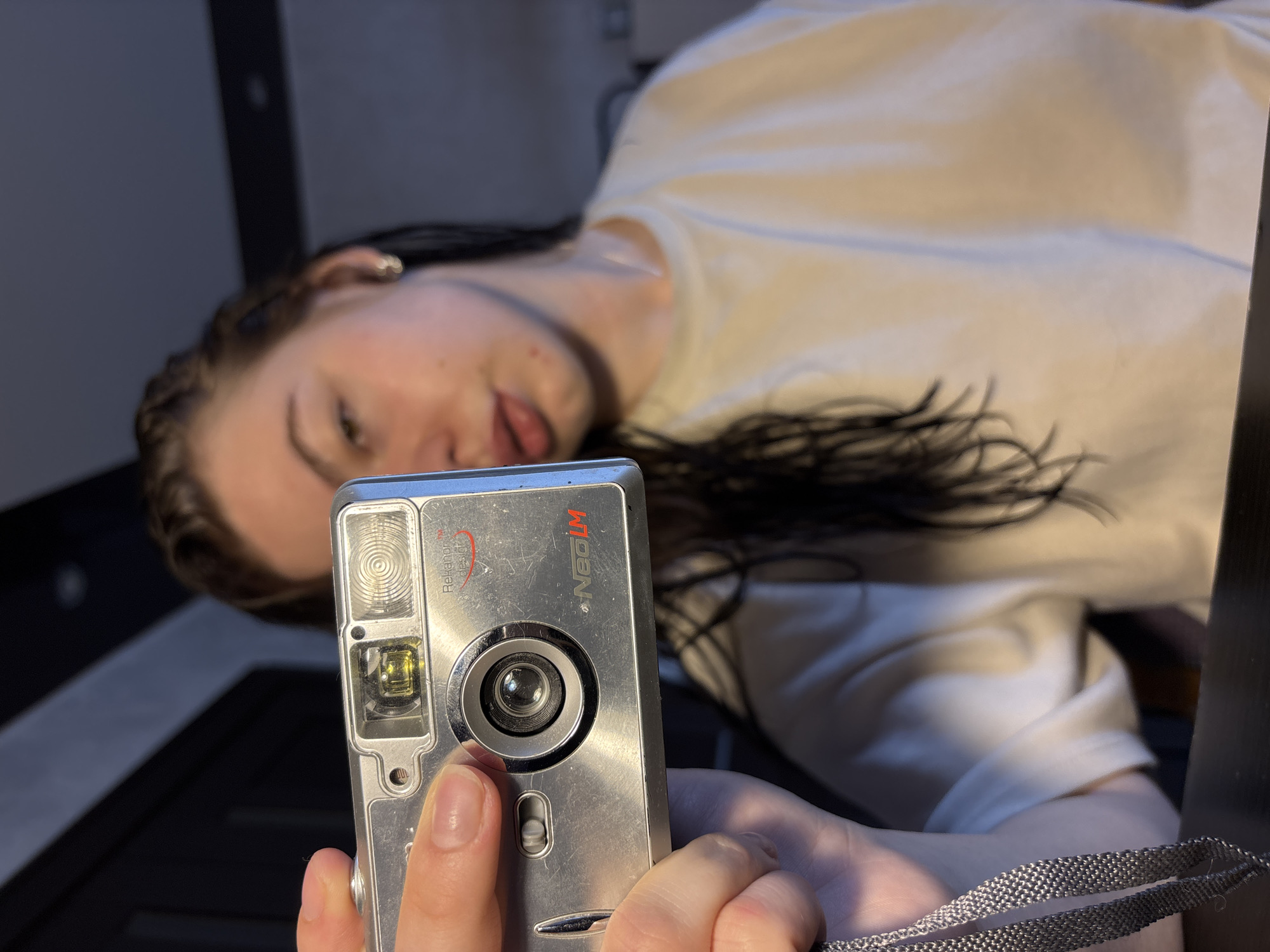 A woman holds a silver NeoLM camera in front of her face, presumably taking a selfie. She wears a white t-shirt. Her long dark hair is visible. The background is blurred, suggesting an indoor location. The camera is the prominent subject in the foreground. There is no information in the image to identify that it was taken in Perm, Russia.
