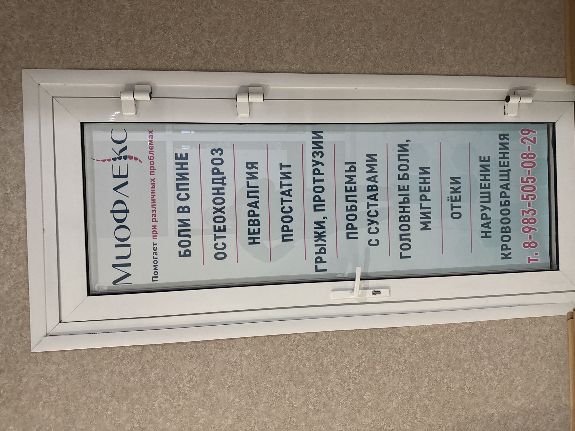 The image shows an advertisement in Russian for a medical clinic, "Miofleks," which treats various problems, including back pain, osteochondrosis, neuralgia, prostatitis, hernias, joint problems, headaches, migraines, swelling, and circulatory disorders. The phone number is 8-983-505-08-29. Based on the phone number prefix and Cyrillic text, it is likely located in Krasnoyarsk, Russia. The advertisement is mounted in a window of a white plastic frame. The wall behind the window appears to be textured and beige.