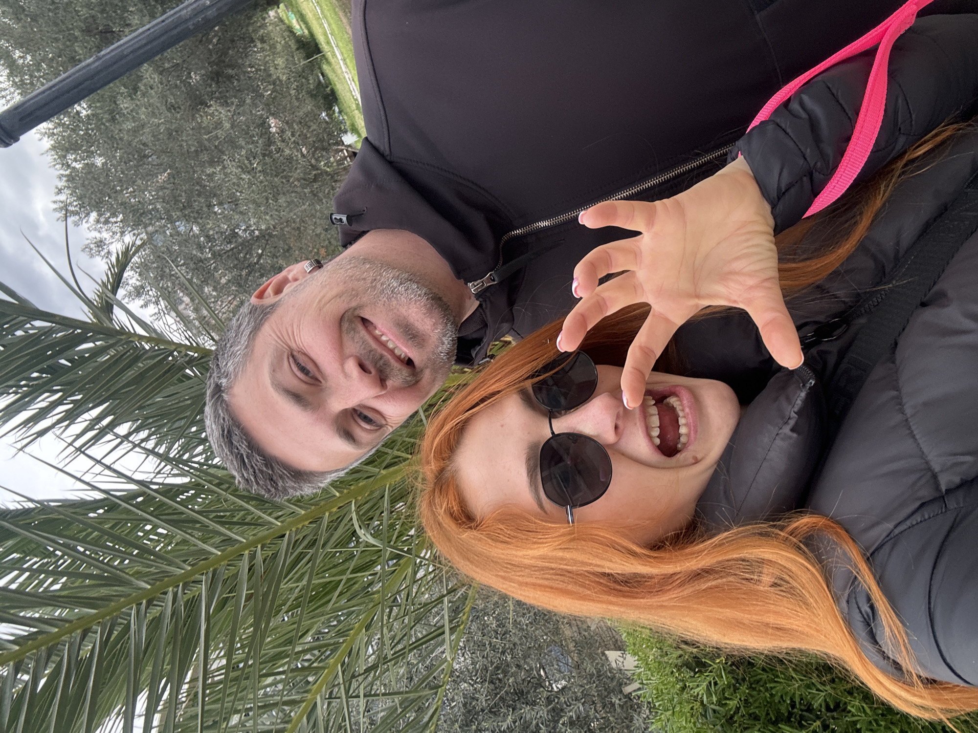 Two people, a man and a woman, are posing for a selfie outdoors. The man, on the left, has short gray hair, a slight beard, and is wearing a black jacket. He is smiling. The woman, on the right, has long red hair and is wearing round sunglasses and a black puffer jacket. She is also smiling and has her hand raised with fingers spread. Palm leaves are visible in the background. The location is Budva, Montenegro.