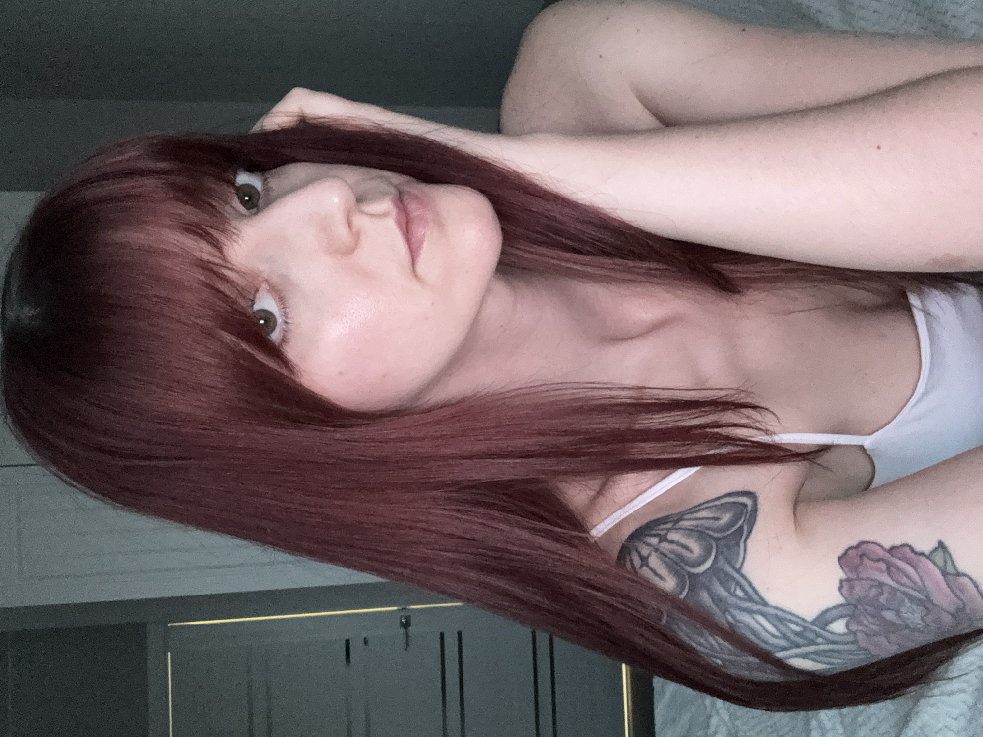 A woman with reddish-brown hair, featuring bangs, is lying down, presumably on a bed. She is fair-skinned and has light brown eyes. She is wearing a white tank top. Her arms are positioned near her head. Tattoos are visible on her upper arm. The background includes a gray wall and a section of what appears to be a dresser or cabinet with a gold trim. There is no evidence to suggest the location is Izhevsk, Russia.