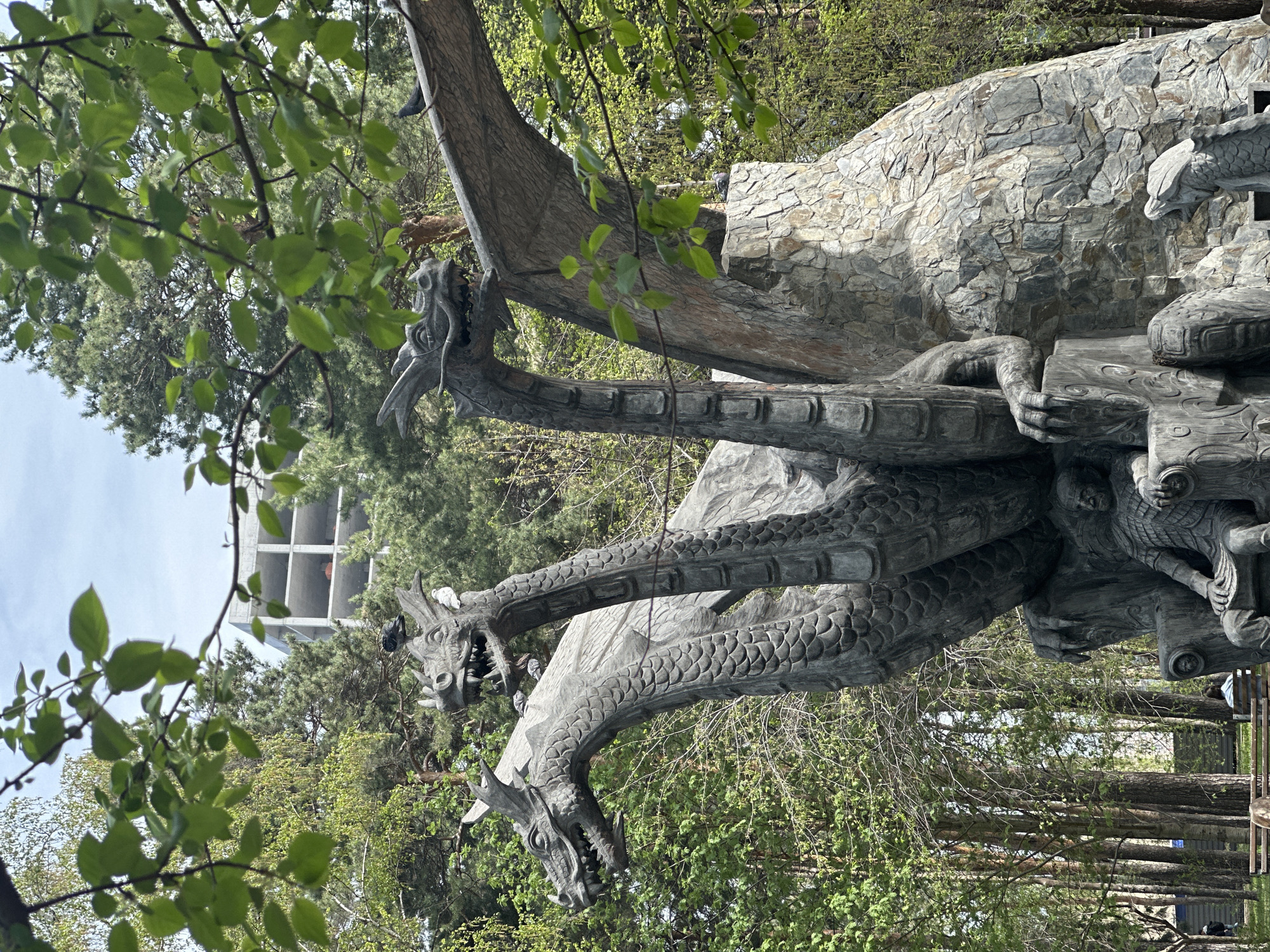 The image shows a stone sculpture of a three-headed dragon in a park setting. The dragon is gray with intricate carvings depicting scales and other details. The sculpture is positioned against a backdrop of trees. A building with a visible concrete structure is visible in the background to the left, indicating a possible urban environment. Based on the information provided, the location is the city of Novosibirsk, Russia.