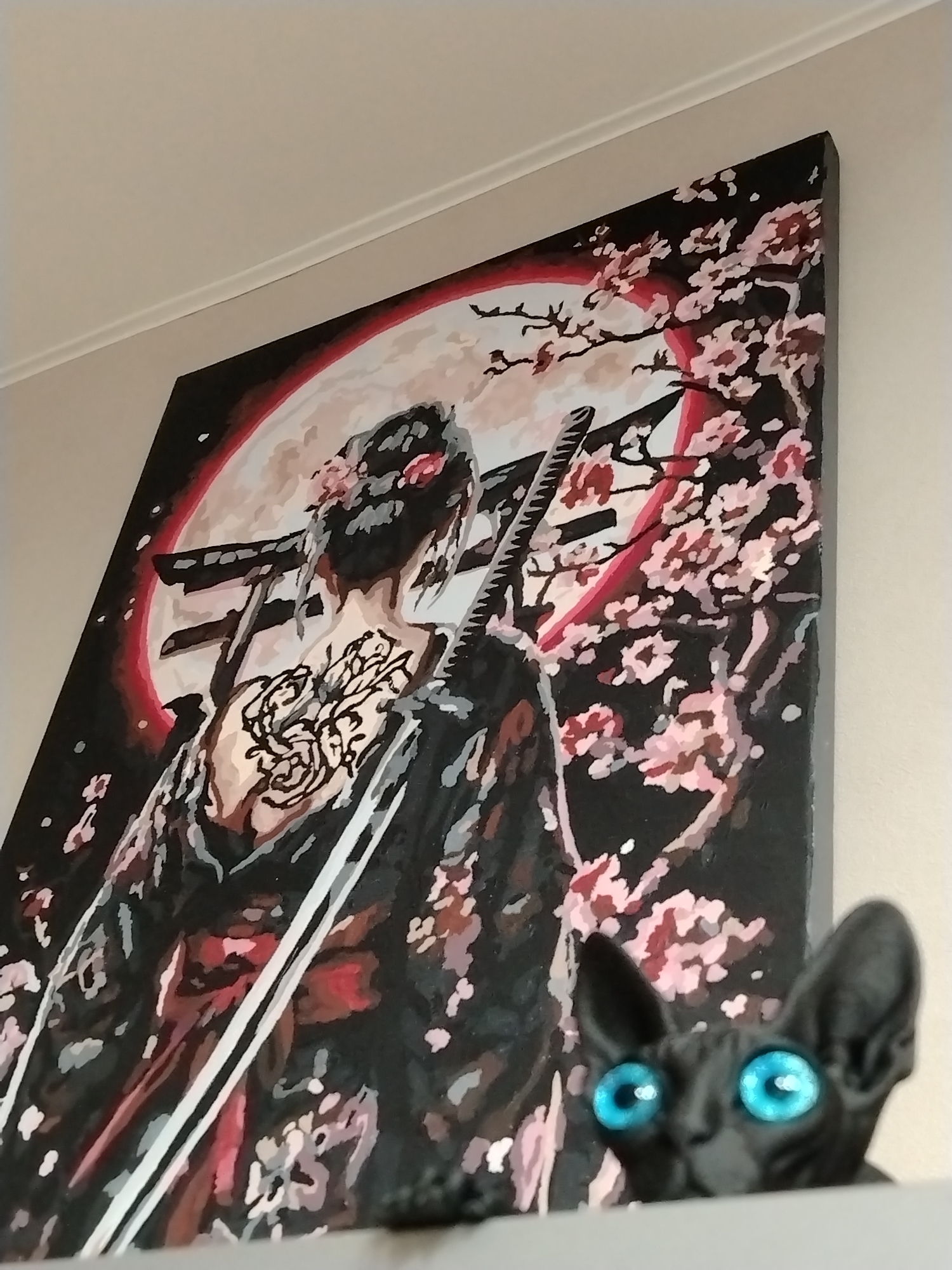 A low-angle shot of a painting on a wall. The painting depicts a woman in a kimono with a tattoo on her back, facing away from the viewer, with a sword and a moon behind her. Cherry blossom branches frame the painting. In the foreground, the head of a black cat statue with bright blue eyes peeks over a shelf or table. The wall is light-colored, with a white molding near the ceiling. This scene is unlikely to be in Izhevsk, Russia, as the style of the painting and statue do not reflect the typical aesthetics of that region.