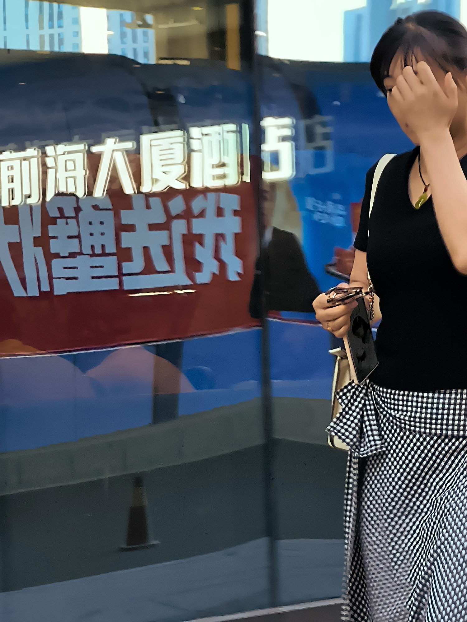 A medium shot captures a woman standing in front of a glass storefront in Ürümqi, China. The storefront displays large, illuminated Chinese characters on a dark blue background, with a red horizontal stripe running across the lower portion of the text. The woman, with dark hair, is wearing a black V-neck top and a black and white checkered skirt with ruffles. She is holding a pair of eyeglasses in her right hand and a dark-colored object, possibly a phone, in her left. Her face is partially obscured as she touches her forehead with her right hand. The environment appears to be an urban setting, with reflections of buildings visible in the glass. A traffic cone is visible in the reflection on the lower left.