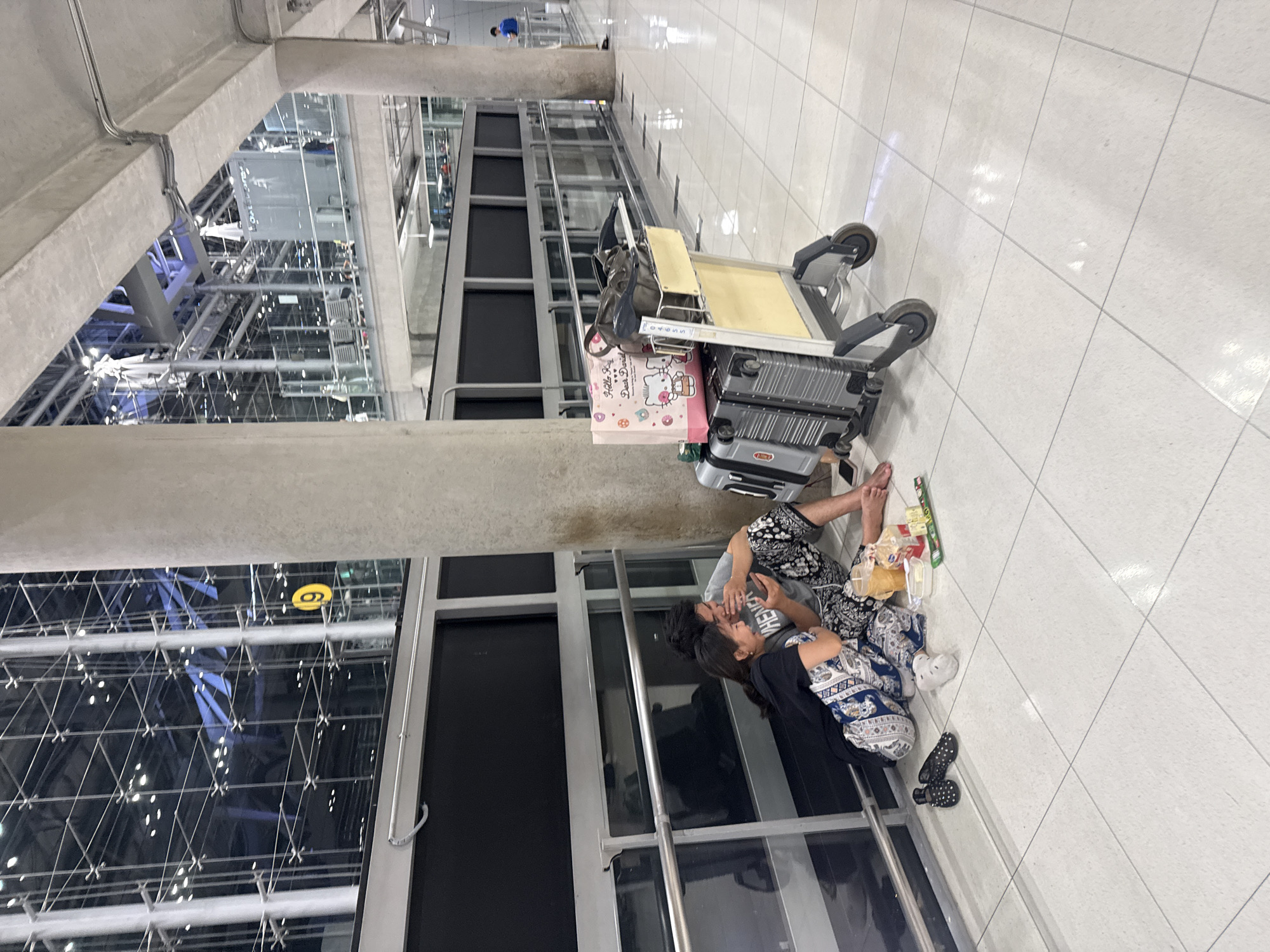 In the vicinity of Nong Prue Subdistrict, Thailand, specifically within an airport terminal, two individuals are seated on the floor near a large pillar. They are positioned on the white tiled floor adjacent to a dark paneled wall with a metal railing. The man, wearing a grey t-shirt and patterned pants, is leaning against the pillar, holding a smartphone and gesturing with his other hand. The woman, dressed in a black t-shirt and matching patterned pants, is seated to his left, also looking at the phone. They appear to be interacting with each other and sharing a moment of amusement, with the woman laughing.
In front of them, on the floor, is a makeshift picnic spread. It includes a plastic container of food, possibly snacks, and a plastic cup with a yellow beverage. Nearby, a pair of black studded flip-flops are placed.
To their right, a luggage cart is loaded with several pieces of luggage. Stacked on the cart are two grey suitcases, one large and one medium-sized, and on top of those is a pink gift bag with a Hello Kitty design. A dark brown bag is also placed on the cart. The overall environment suggests a waiting area in a modern airport, with a high ceiling and expansive windows revealing the structure of the terminal. The lighting is ambient, contributing to a somewhat dim atmosphere within the terminal.