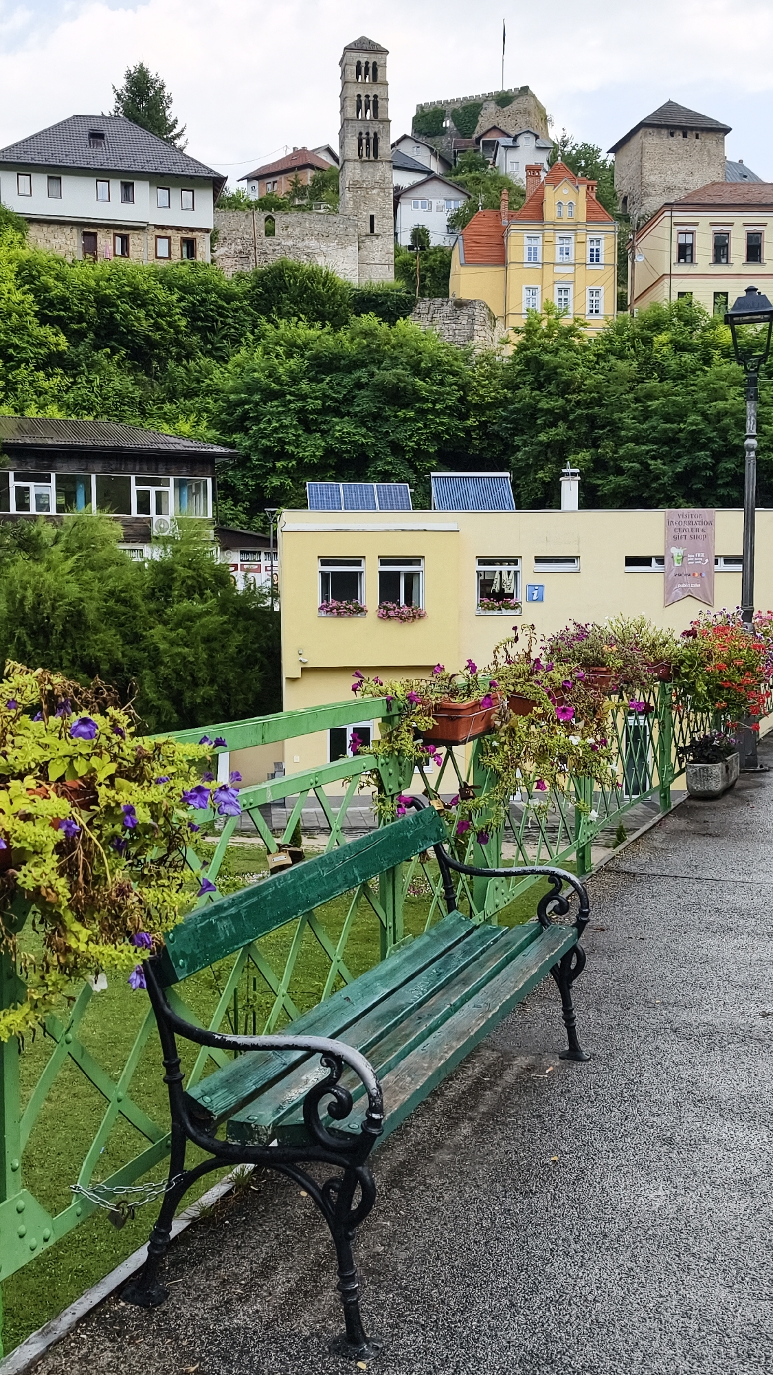 The image shows a picturesque view of Jajce, Bosnia and Herzegovina, featuring the historic Old Town and the Pliva River. In the foreground, a weathered green park bench with ornate black cast-iron legs sits on a paved walkway. The bench is adorned with hanging planters filled with vibrant purple and red flowers, adding a touch of color to the scene.
Behind the bench, a bright yellow building with solar panels on its roof is visible. This building appears to be a visitor information center, as indicated by a banner on its facade. The banner reads "VISITOR INFORMATION CENTER & GIFT SHOP." The building has several windows, some with flower boxes, and a distinctive number "7" above one of them.
Further up the hill, the iconic towers and walls of Jajce's fortress dominate the skyline. A prominent stone tower, likely part of a historical church or minaret, stands tall amidst the lush green foliage that covers the steep hills. Various other buildings, painted in different colors and architectural styles, are nestled on the slopes, showcasing the unique charm of the city. The overall atmosphere is serene and inviting, with a sense of history and natural beauty coexisting. The sky is overcast, suggesting a slightly humid or rainy day.