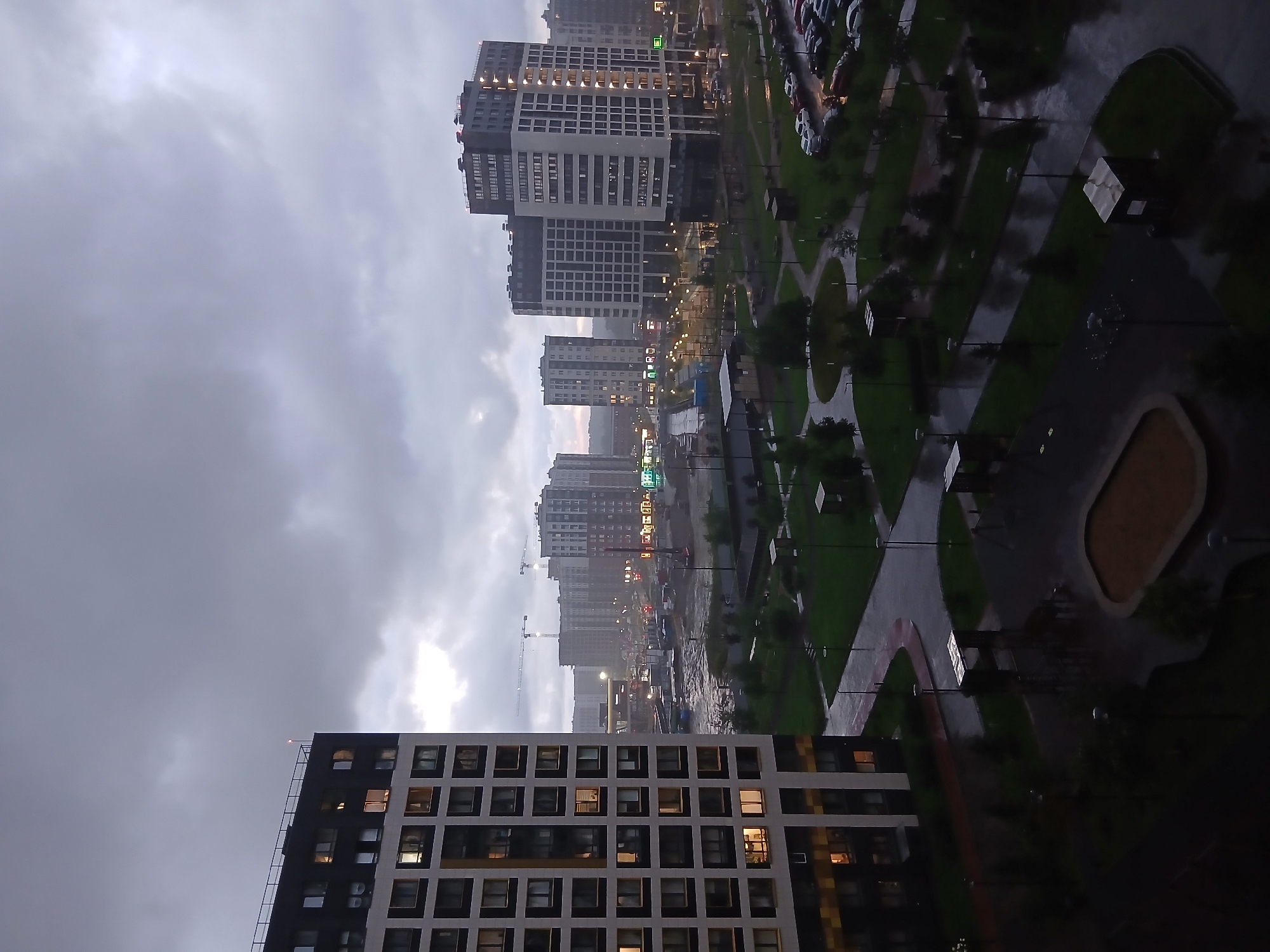 The image depicts a view of the city of Murino, Russia, on a cloudy and possibly rainy day. In the foreground, a tall, modern apartment building with a grid-like pattern of windows dominates the left side of the frame. The building has sections of dark and light colored facades, and some windows are lit from within. Across from this building, a street with parked cars is visible.
Further into the background, more residential buildings, also modern in design, are spread across the landscape. Some of these buildings are under construction, indicated by cranes and scaffolding. The overall atmosphere is somber, with overcast skies casting a diffused light over the scene. The ground appears wet in places, suggesting recent or ongoing precipitation. The urban environment shows a mix of completed and developing residential areas, typical of a growing city. There are no people clearly visible in the image, nor any specific actions or interactions taking place. The focus is on the architecture and the overall urban setting under a heavy sky.