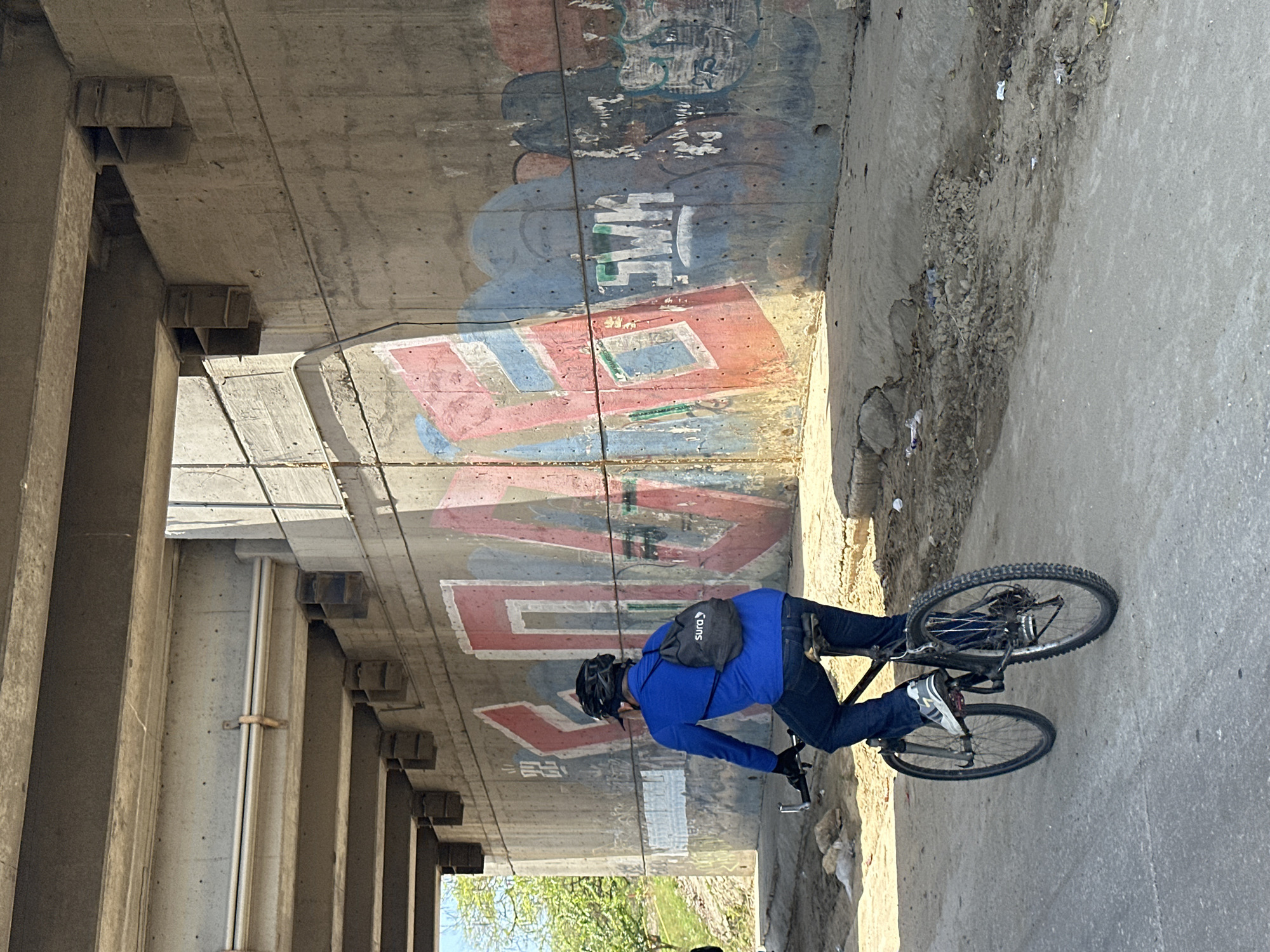 In Barranquilla, Colombia, under a concrete overpass, a person rides a bicycle on a paved road. The overpass is supported by thick concrete columns and beams, with visible rebar and concrete construction details. The wall beneath the overpass is extensively covered in graffiti, featuring large, stylized letters in red, blue, and white, with some areas of faded or partially obscured tags.
The cyclist, seen from behind, wears a blue long-sleeved shirt, dark pants, and a black helmet. A small, dark backpack with a white logo that reads "SURA" is slung over their shoulders. The cyclist is on a dark-colored bicycle with silver handlebars and black tires. Their right foot is on a pedal.
The road surface is asphalt, with a sandy, uneven area to the right of the cyclist, littered with small rocks and debris. The environment appears somewhat unkempt, with a general sense of urban infrastructure and decay. Sunlight casts shadows from the overpass onto the wall and the road, indicating it is daytime. Trees with green foliage are visible in the background on the left, suggesting a vegetated area beyond the overpass.