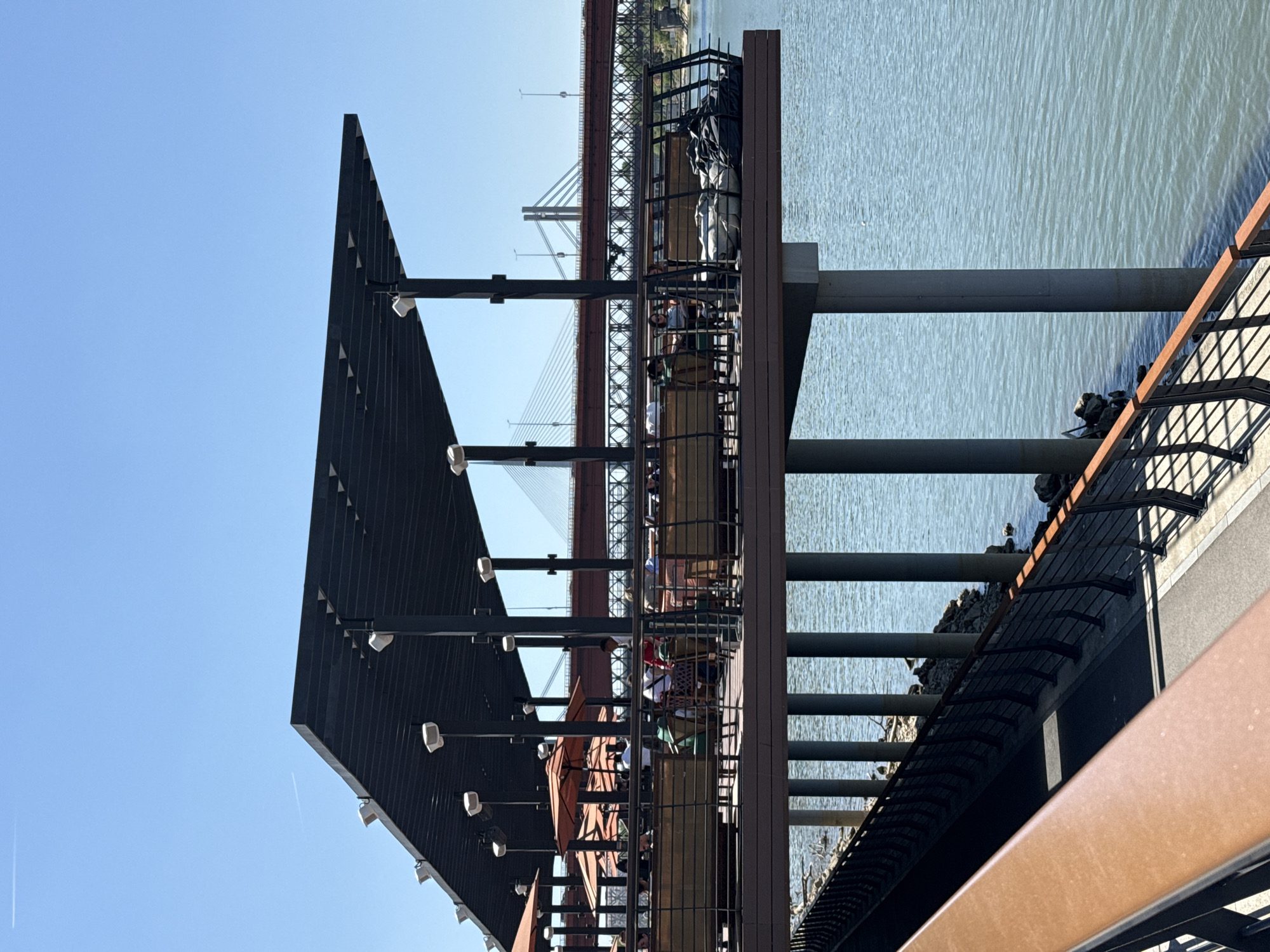 The image shows a waterfront restaurant in Belgrade, Serbia, with the Branko's Bridge in the background. The restaurant has a modern architectural design with a dark metal canopy and outdoor seating. Patrons are visible at the tables. The restaurant is elevated on pillars above the water. The Danube River is to the right of the restaurant. A paved walkway with a railing is in the foreground. The sky is clear and blue.