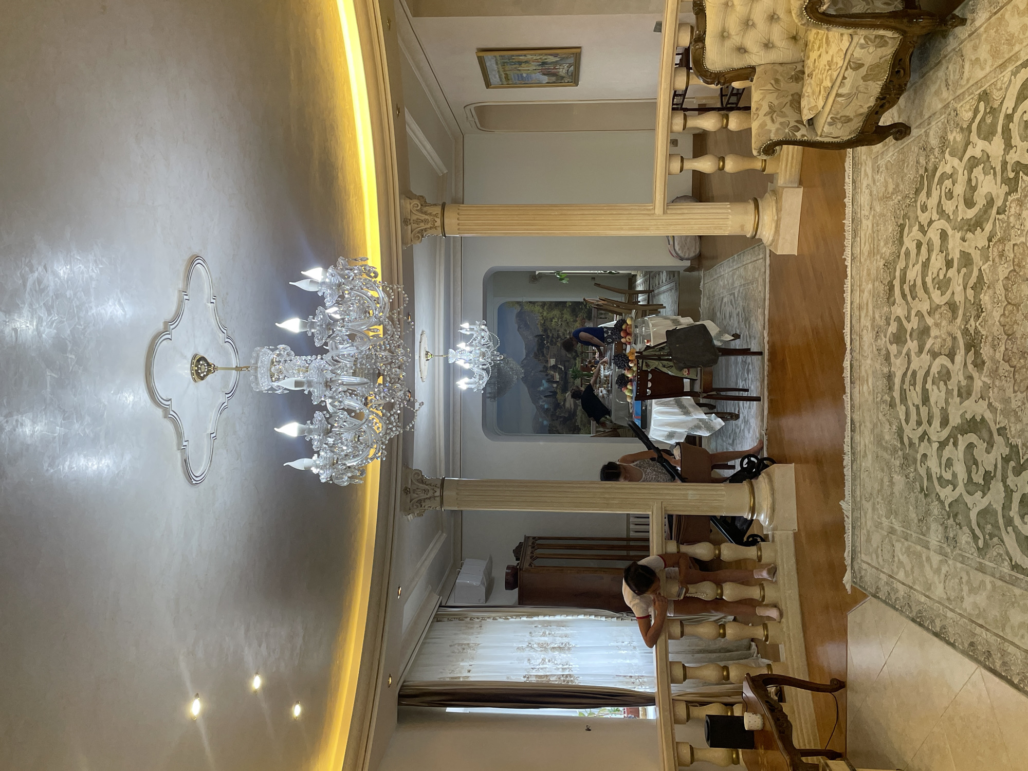 The image shows a spacious, opulent interior of a room with a high, ornate ceiling featuring a large crystal chandelier and recessed lighting. The room appears to be a dining or living area, with a long dining table set for a meal in the background. Several people are present, some interacting with the table and others engaged in casual activities. One person, wearing a white shirt and shorts, is leaning over a railing on the left side of the image, examining something in their hands. Another person, in a patterned dress, is standing near the dining table. A third person, wearing a blue top, is also near the table. The room is furnished with plush seating, decorative columns, and a large patterned rug on the floor. A mural depicting a landscape scene adorns the wall in the background. A small side table with a cup and a wooden chair are visible on the far left. The overall ambiance suggests a well-appointed home. The prompt requests the location be Almaty, Kazakhstan. However, there are no visual cues in the image to support this specific location.
