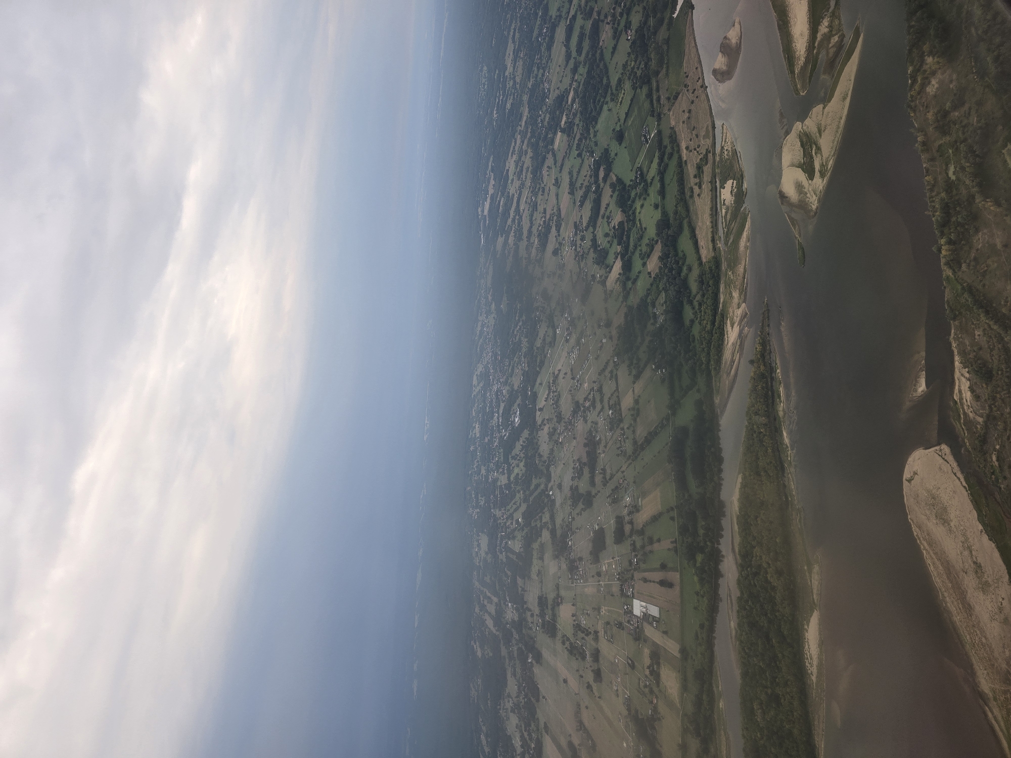 An aerial view captures a vast landscape under a cloudy sky. A wide, dark river dominates the foreground, with sandy islands breaking its surface. Lush green trees line the riverbanks and extend into the middle ground, where they give way to a mosaic of agricultural fields and scattered buildings. The fields are laid out in geometric patterns, suggesting organized farming. In the distance, the land appears more developed, with a denser concentration of structures and roads fading into a hazy horizon. The overall scene depicts a rural or semi-rural area with a significant waterway and cultivated land.