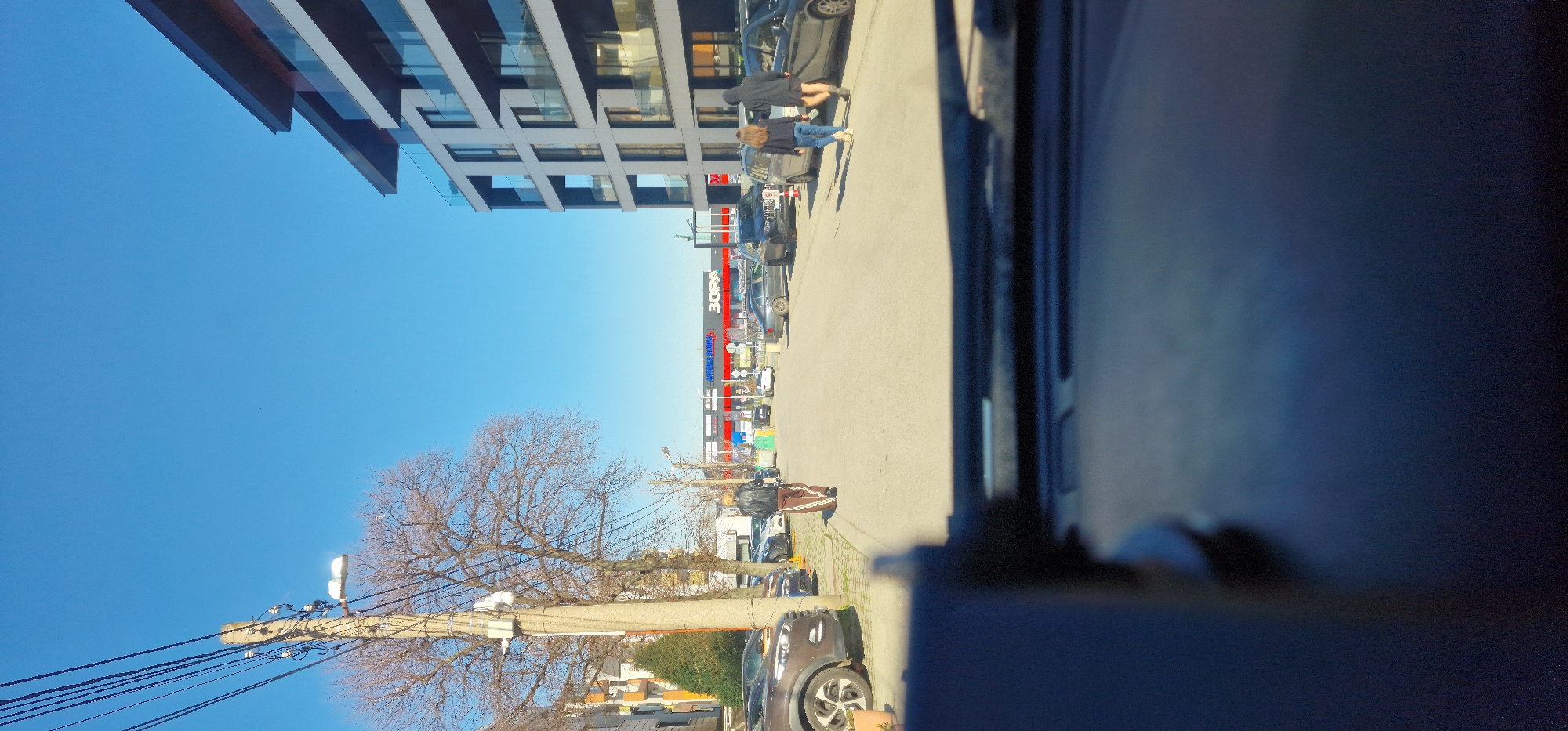 The image shows a street scene in Sofia, Bulgaria, with a clear blue sky above. A multi-story modern building with numerous windows stands on one side of the street, casting a shadow on the pavement. A supermarket named "Zora" is also visible. Several parked cars line the street. Two people are walking on the sidewalk. A leafless tree and a utility pole with multiple wires are on the opposite side of the street. The photograph appears to be taken from inside a vehicle, with a portion of the vehicle's interior visible in the foreground.