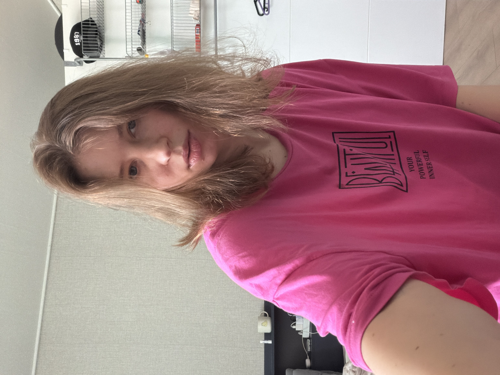 A young woman with shoulder-length blonde hair is positioned in the foreground, looking directly at the camera. She is wearing a bright pink t-shirt with black text on it that appears to say "BEAUTIFUL" above the words "YOUR POWERFUL INNER SELF." The background features a light-colored textured wall, a shelf with a mug and a tube of lip balm, and a white closet with wire shelving holding a black baseball cap with "1987" on it. A dark blue headboard or shelf is visible behind the woman. The overall setting suggests an indoor, possibly domestic, environment. The image was captured in Dongducheon-si, South Korea.