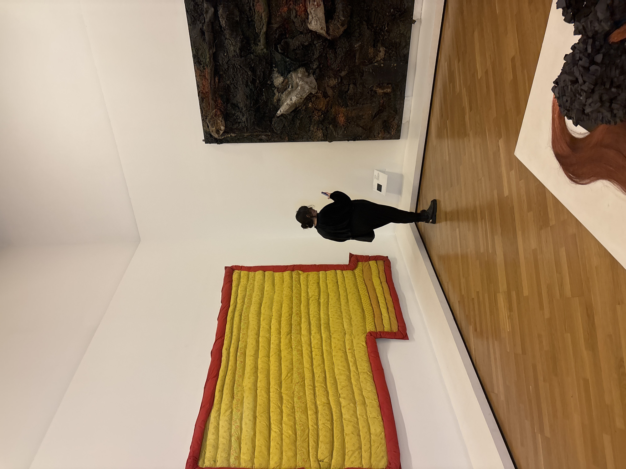 A person in dark clothing stands looking at artwork in a gallery with white walls and wood flooring. The artwork includes a dark, textured piece above the person, a yellow and red quilted piece to the left, and a white piece with black and brown elements to the right. The person is holding a device, likely a phone, and is oriented towards the dark, textured piece. The scene likely takes place in a contemporary art museum. The location in São Paulo, Brazil is not discernible from the image.