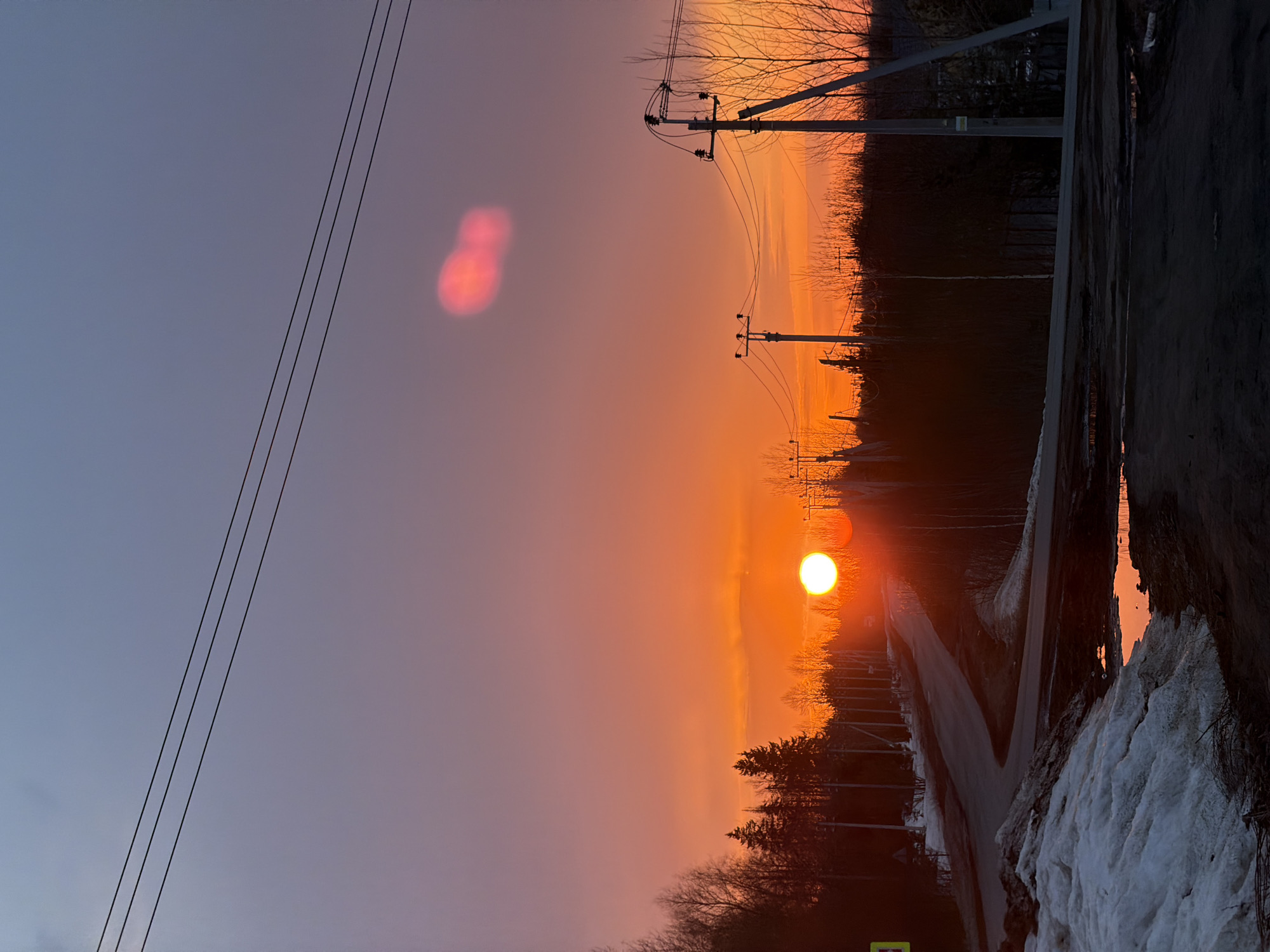 The image captures a vivid sunset in Izhevsk, Russia. The sky transitions from a muted blue-grey to a fiery orange and red near the horizon. Power lines are visible on the left side of the frame, and utility poles line the road leading towards the setting sun. The sun itself is a bright, spherical orb partially obscured by trees and haze. A road runs through the center of the scene, flanked by trees and residual snow. Patches of snow and ice are visible on the right side of the image, near the edge of the road, with reflections of the sunset in standing water. The scene is generally devoid of people.