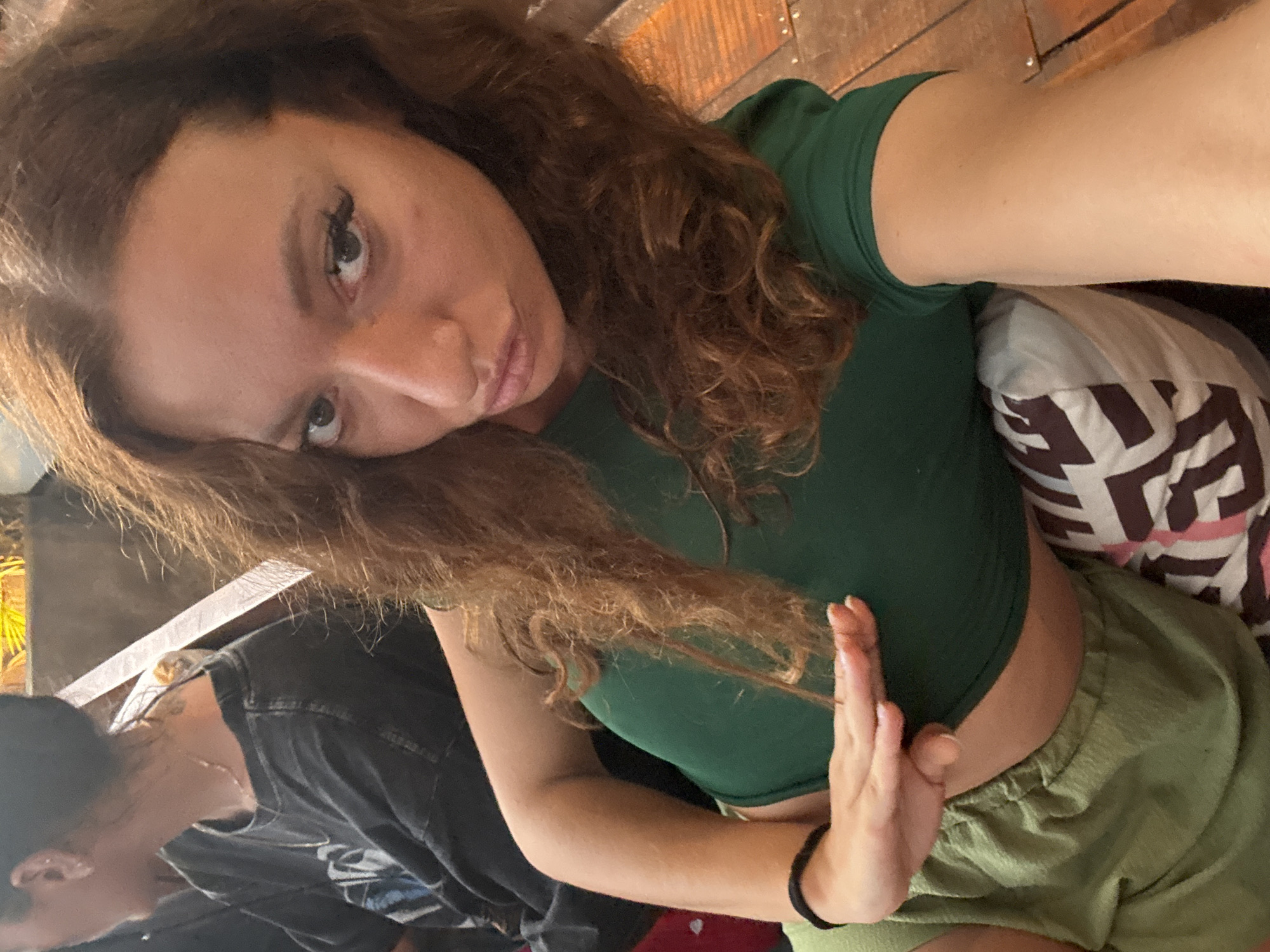A woman with long, wavy brown hair and light skin is posing for a selfie. She is wearing a green crop top and green shorts. Her left hand is raised in front of her torso. Behind her, there is a patterned pillow. To the left of the woman, there is another person wearing a dark shirt. The background appears to be a wooden surface. The location is specified as Hà Giang 1 Ward, Vietnam.