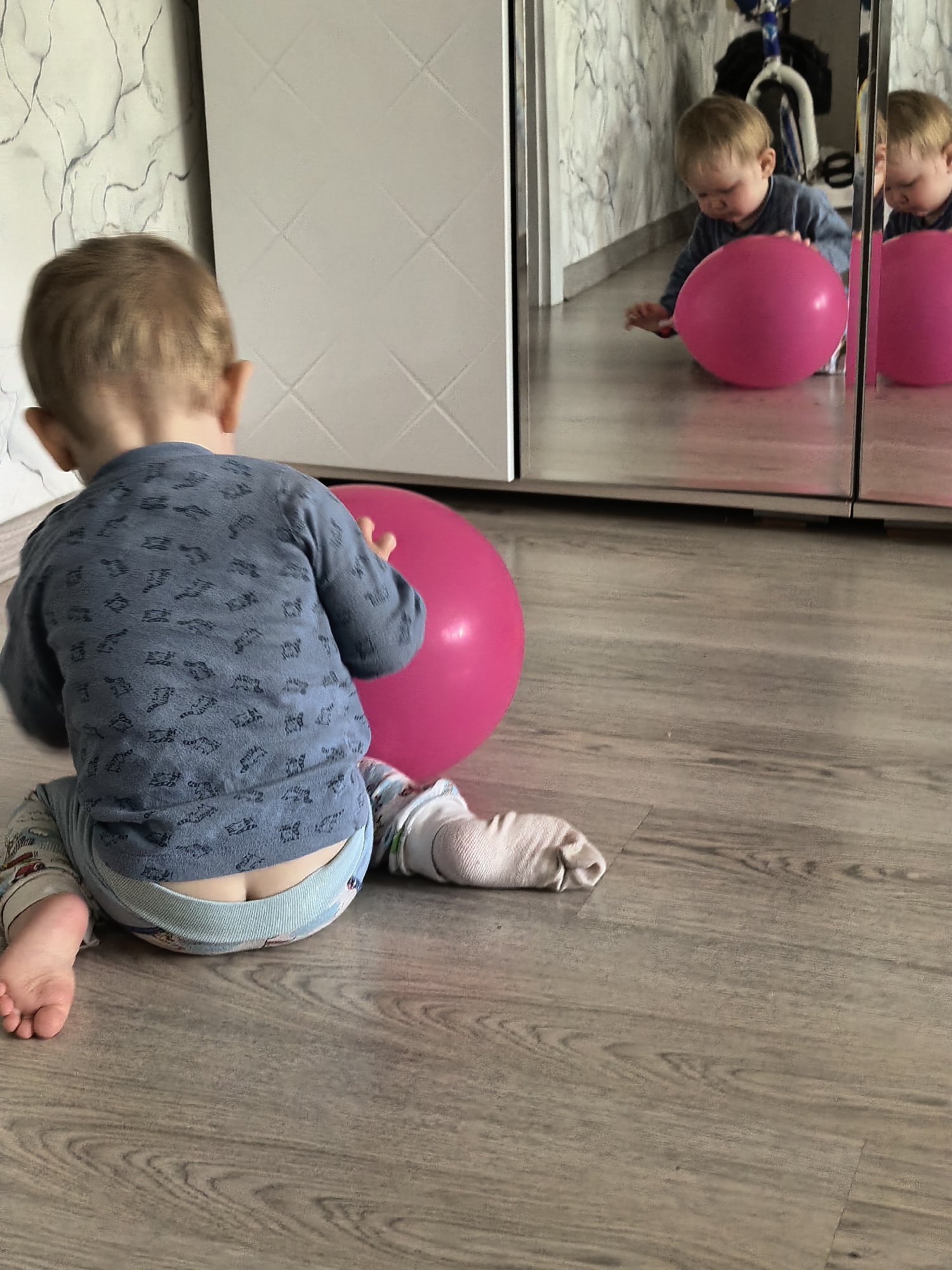 An indoor scene depicts a young toddler sitting on a light wooden floor, viewed from behind. The toddler has light brown hair and is wearing a blue long-sleeved top with dark patterns, light-colored patterned pants, and a diaper peeking out above the waistband. Their left foot is bare, while the right foot is covered by a light-colored, possibly knitted, sock or bootie.
The toddler is focused on a vibrant pink exercise ball situated to their right. In front of the toddler and to the right, a large mirrored surface, likely part of a wardrobe or cabinet, reflects the scene. The reflection shows the front of the toddler, appearing to reach out or look intently at the pink ball. Behind the reflection of the toddler, a blue bicycle is partially visible. The background wall to the left of the mirror features a white patterned design with dark, swirling lines. To the left of the actual toddler, there's a plain white cabinet or wall with a subtle diamond pattern.
The lighting suggests daytime, casting a soft, even glow across the room. The overall impression is one of a child engaged in play or exploration within a home environment. No text is visible in the image.