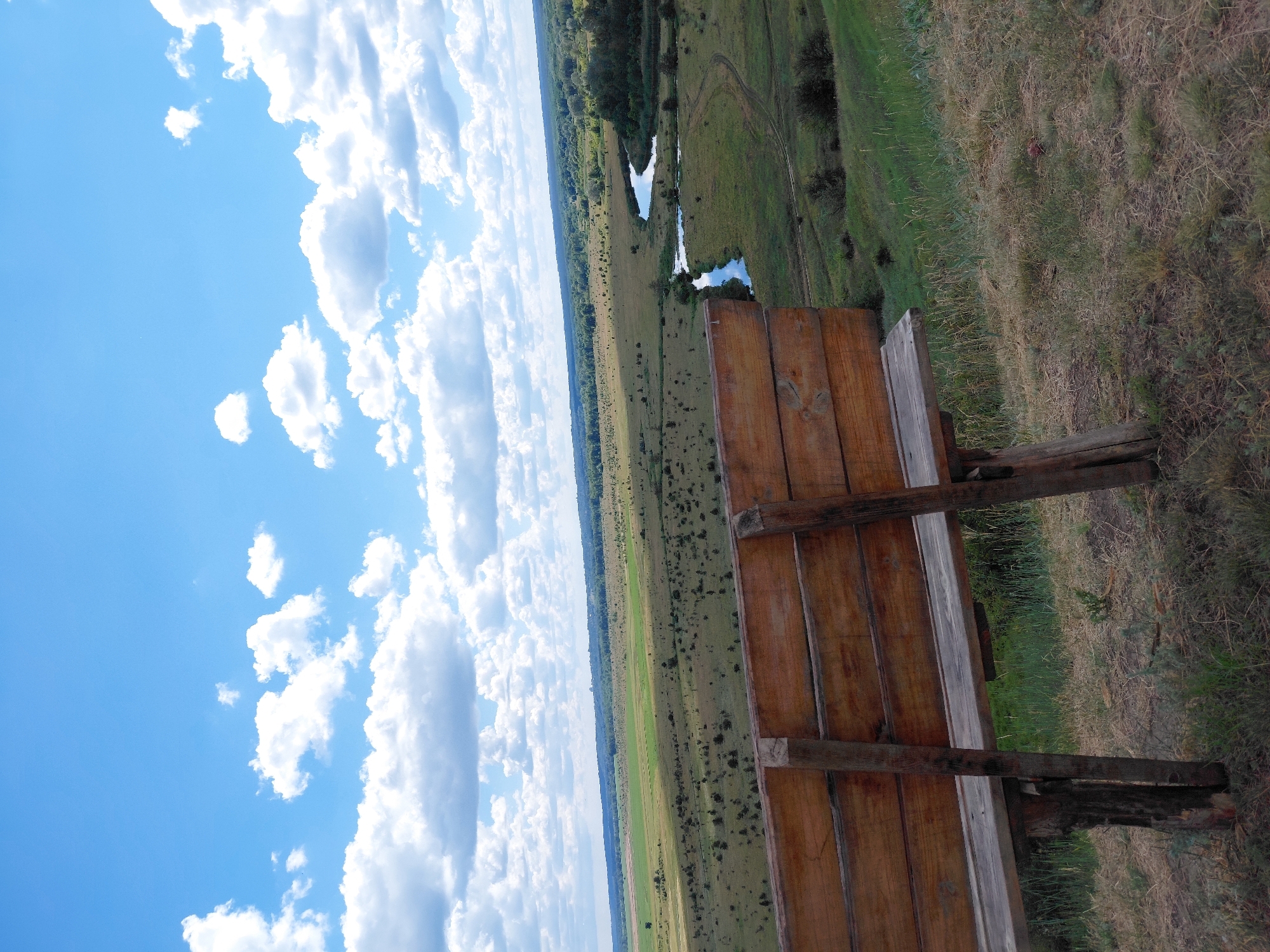 The image depicts a wooden bench positioned in the foreground, facing a vast landscape. The bench is constructed from rough-hewn wooden planks and supported by sturdy wooden posts. Behind the bench, a wide expanse of green fields stretches towards the horizon, dotted with scattered trees and shrubs. A winding river meanders through the landscape, reflecting the blue sky and fluffy white clouds above. The sky is a vibrant blue, filled with cumulus clouds. The overall scene conveys a sense of natural beauty and tranquility. The location is identified as Ustyvytsia, Ukraine.