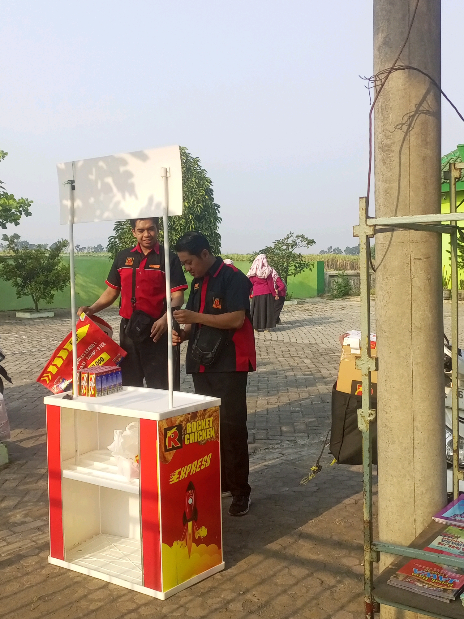 In Gurah, Indonesia, two men in matching red and black uniforms stand at a portable food stand. The stand is white and red, decorated with the branding "Rocket Chicken Express" and a rocket graphic. One man holds a red and yellow promotional sign, while the other appears to be organizing items on the counter. A white canopy is erected above the stand. In the background, trees, a paved area, and a building are visible. Several women in headscarves are also seen in the distance, walking across the paved area. The scene takes place outdoors during daylight hours, with clear skies.