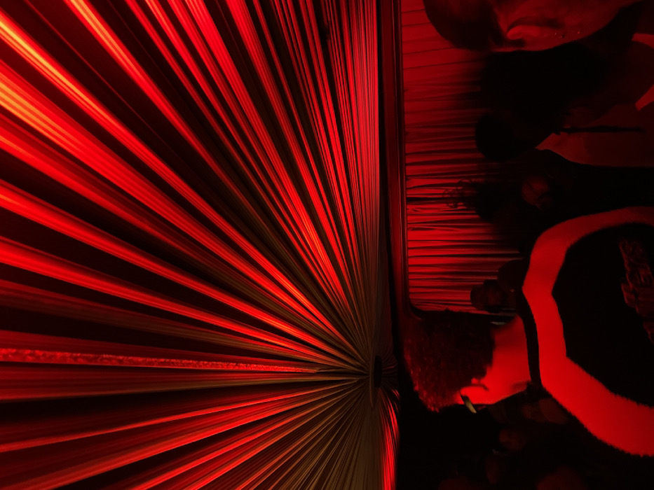 The scene depicts an indoor event space dominated by intense red lighting. The ceiling is adorned with red fabric draped in a radial pattern, emanating from a central circular fixture. Below this ceiling, a dark horizontal band separates the upper structure from a red curtain that covers the entire background. Several individuals are present, visible primarily as silhouettes or partially illuminated by the crimson light. In the left foreground, a person with dark, curly hair is seen from behind, wearing a dark garment with prominent light-colored sections on the shoulders. To the right, a portion of another individual's head and ear is visible in profile. Additional indistinct figures occupy the midground and background. No specific identifying markers are visible within the image to determine a precise location within the city of Paris, France.