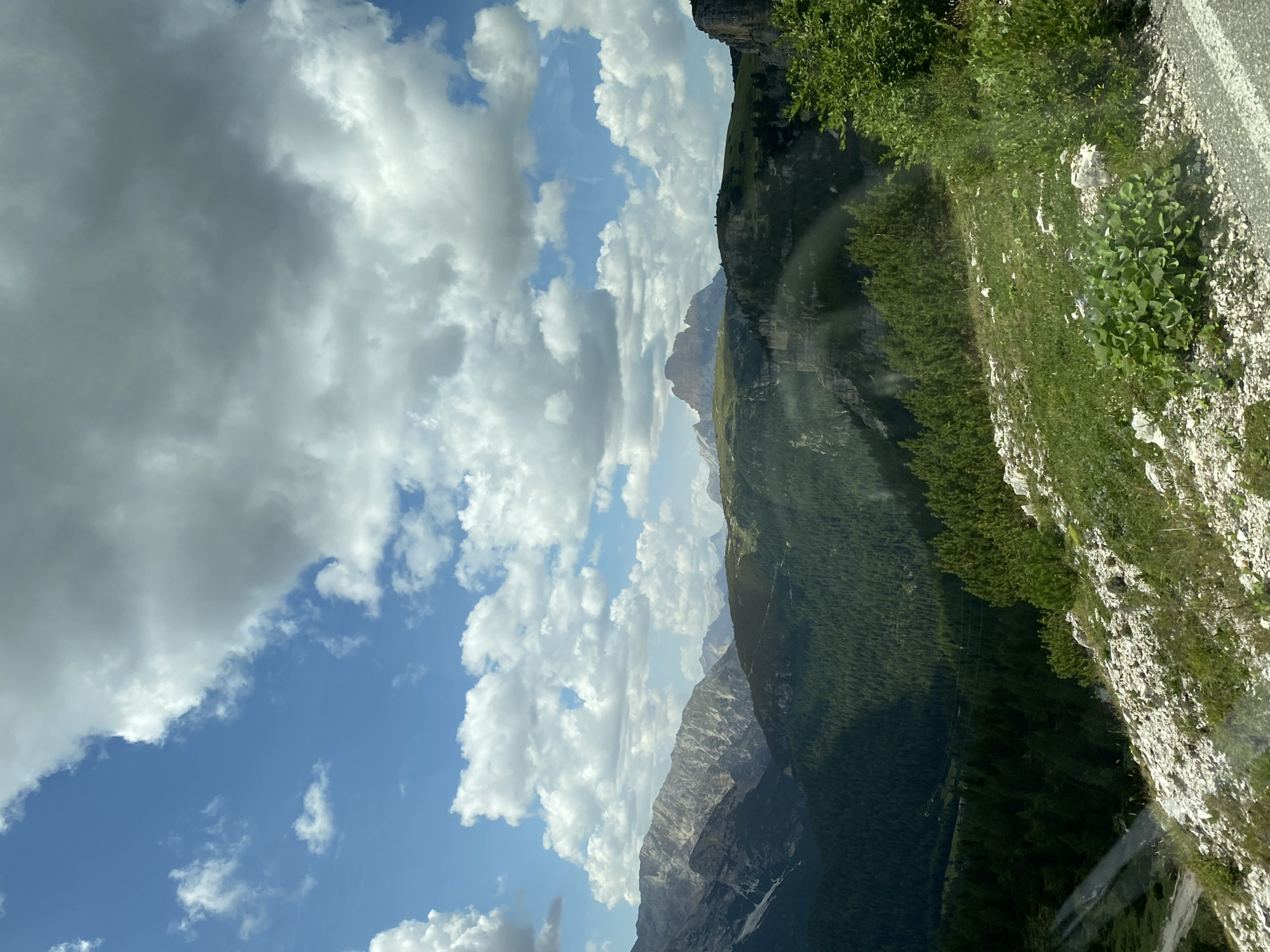 This is a high-angle, long-shot photograph of a mountainous landscape in Auronzo di Cadore, Italy. The sky is a vibrant blue, filled with a mix of fluffy white cumulus clouds and darker, more ominous storm clouds. The terrain below is rugged and dramatic. Jagged, rocky mountain peaks, some with patches of snow, dominate the background. Below these peaks, a dense, dark green coniferous forest blankets the slopes, creating a stark contrast with the lighter, rocky terrain. In the foreground, a steep, grassy hillside is covered in small white rocks and scattered green vegetation. The scene appears to be captured from a moving vehicle, as evidenced by the slight distortion and light streaks across the lower portion of the image, suggesting it was taken from a car window. There are no people or animals visible in the frame. The overall impression is one of vast, untamed natural beauty under a dynamic sky.