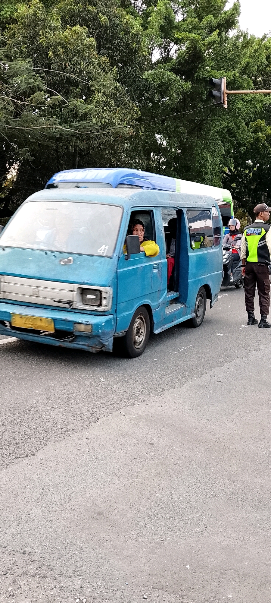A blue van is stopped at a traffic light in Depok, Indonesia. The van has passengers inside and the driver, a woman wearing a yellow shirt, is looking out the window. A traffic police officer in a reflective vest and dark pants stands on the side of the road. A motorcyclist is behind the van. The background consists of trees and a traffic light. The road is paved and appears to be in good condition.