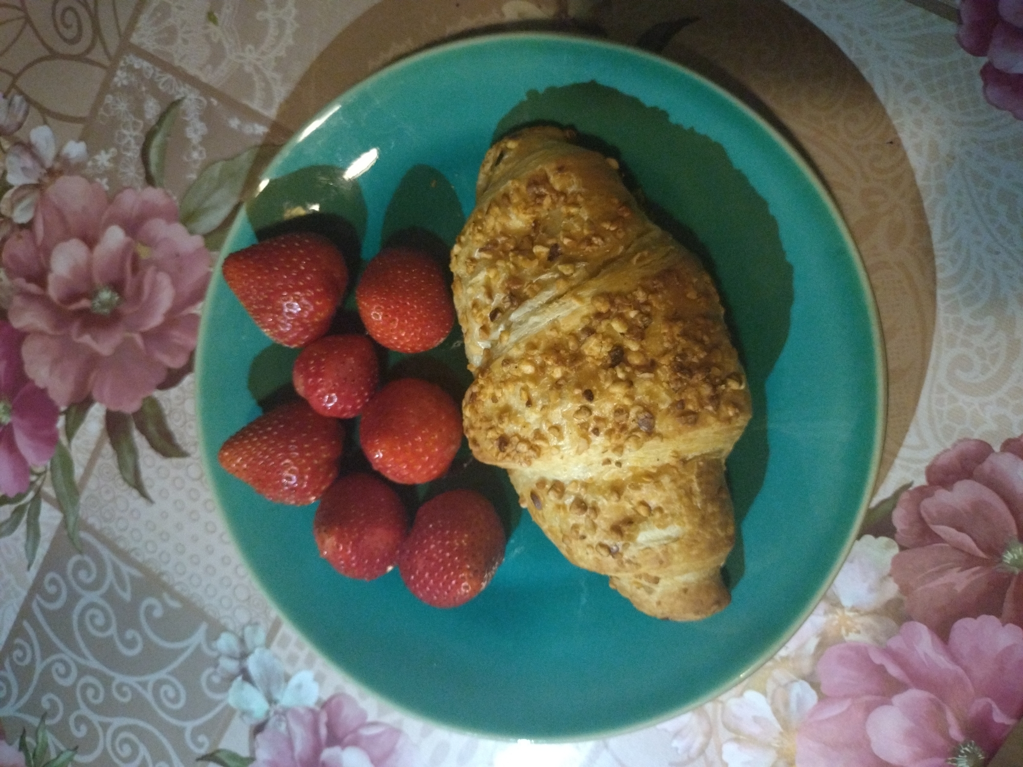 The image displays a close-up, top-down view of a meal, likely breakfast or a snack, featuring a croissant and fresh strawberries. The food items are arranged on a round, teal-colored ceramic plate, which rests on a decorative, patterned tablecloth.
On the right side of the plate is a large, golden-brown croissant. It has a flaky, baked texture and is generously topped with what appears to be chopped nuts, possibly almonds or hazelnuts, giving it an appealing crunchy surface.
To the left, filling the remaining space on the plate, are at least seven bright red strawberries. They appear fresh and ripe, with some showing a slight sheen, suggesting they are juicy.
The plate is placed on a tablecloth with an elaborate pattern. The pattern features large, soft pink flowers and smaller white floral elements, accompanied by green leaves. Interspersed among the floral designs are beige or light brown swirling motifs on a lighter background, creating a rich and warm aesthetic.
No people are visible in the image, and there is no discernible text. The lighting suggests an indoor setting during the daytime, perhaps a casual breakfast or brunch moment. Given the context, the scene is set in Stara Zagora, Bulgaria.