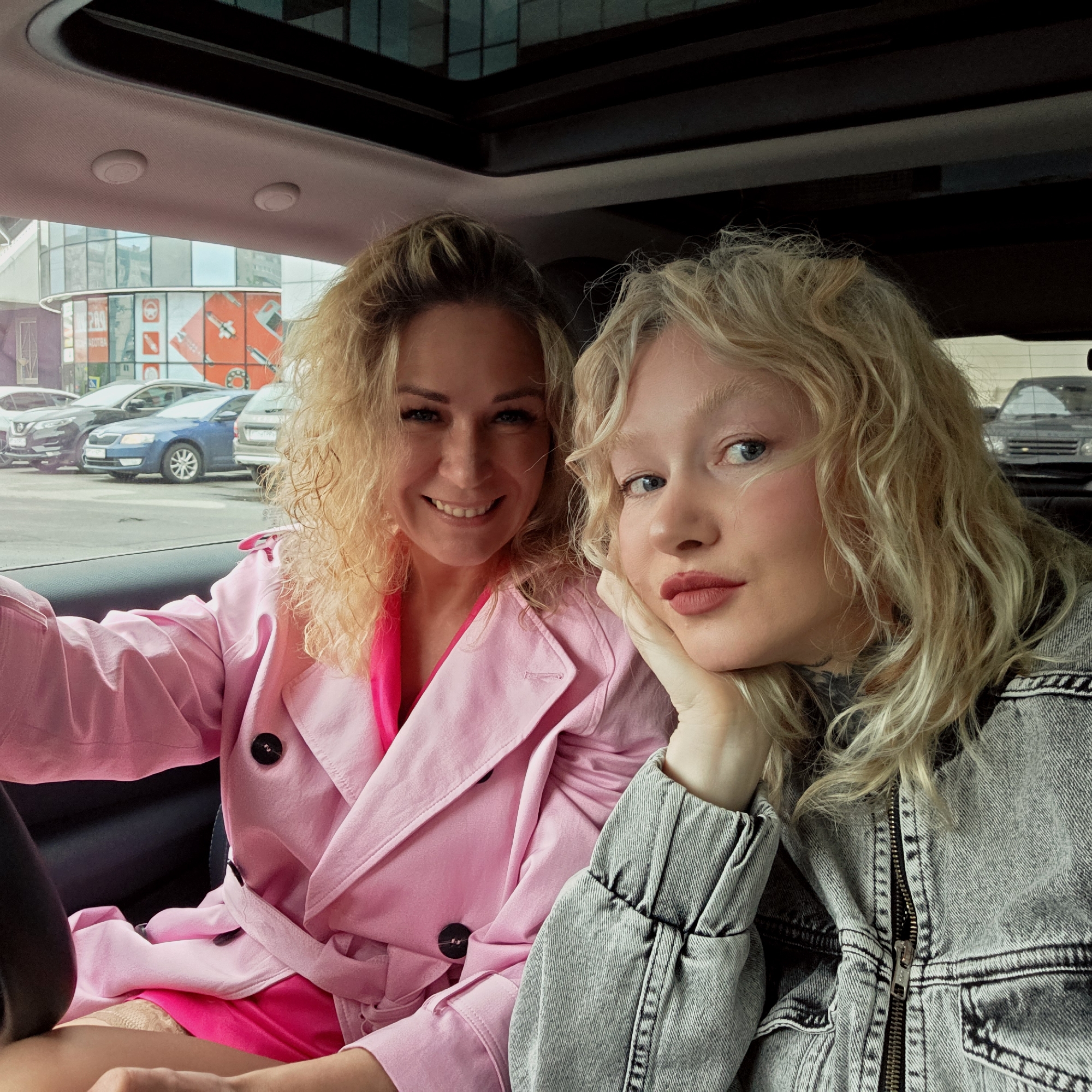 Two blonde women are sitting in a car, possibly parked near a building with advertisements on it, as seen through the car window. The building's advertisement features imagery of car parts and is labeled in Russian. The background also shows multiple cars parked outside the building. The location appears to be Saint Petersburg, Russia, based on the architectural style of the building visible through the car window, as well as the Cyrillic lettering on the advertisements.
