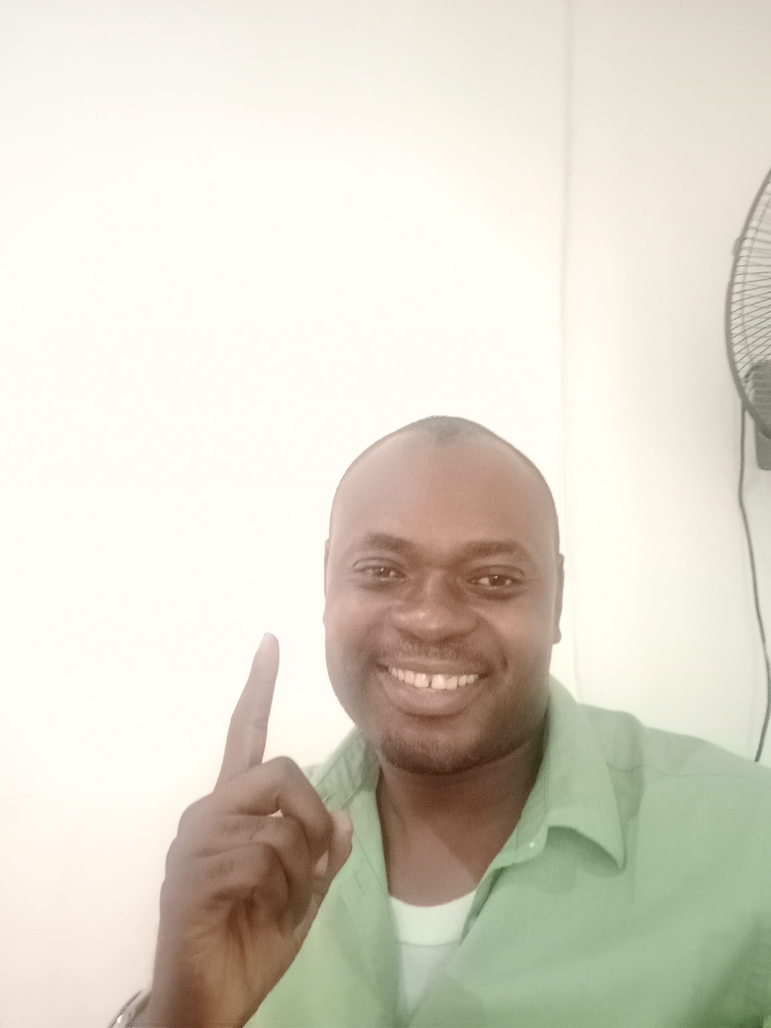 A man with dark skin, a receding hairline, and a friendly, broad smile is featured in a portrait-style image. He is wearing a light green collared shirt over a white undershirt. His left hand is raised with the index finger pointing upwards, and a faint light-colored wristband or watch is visible on his wrist.
The man's expression is joyful, and he appears to be posing for the camera. The setting is indoors, with a plain, light-colored (likely white or off-white) wall forming the background. A corner of the room can be subtly seen behind him. In the upper right portion of the image, the dark protective grate of a ceiling fan is partially visible. The lighting is bright and consistent with an indoor environment. No specific time of day or weather conditions can be inferred from the image, and no text is visible. The location context provided is Gwarinpa, Nigeria.