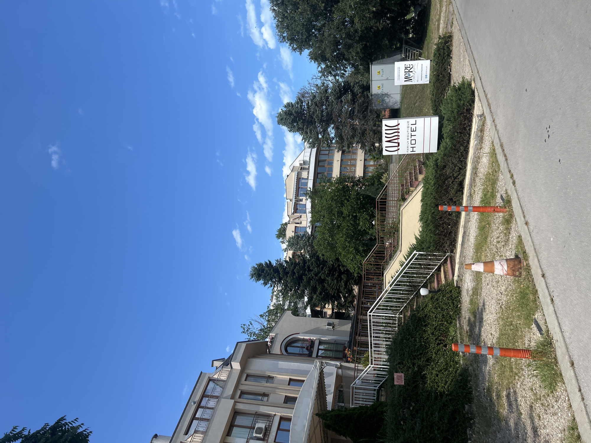 This is a vertically oriented, eye-level photograph of a street scene in Varna, Bulgaria. The day is clear and sunny with scattered white clouds in a bright blue sky. A paved road runs along the right side of the frame, bordered by a strip of gravel and then a neatly trimmed green hedge.
In the foreground, a white traffic cone with orange stripes is placed on the gravel. Behind the hedge, a white staircase with a metal railing leads up towards a building. Further back, a building with a modern design, featuring multiple balconies and large windows, is visible. Another taller building with a curved facade can be seen behind it, partially obscured by lush green trees.
Two signs are prominently displayed on the left side of the road. The closer sign has "CLASSIC FAMILY BOUTIQUE HOTEL" written in dark text on a white background. Behind this, another sign reads "MORE" and displays a phone number and website address. In the distance, another large billboard-like structure is visible.
The overall impression is one of a well-maintained urban environment on a pleasant day. The presence of the hotel signs suggests this is an area with accommodation and possibly commercial establishments. The traffic cones indicate some ongoing work or a temporary obstruction. The scene appears calm and orderly.